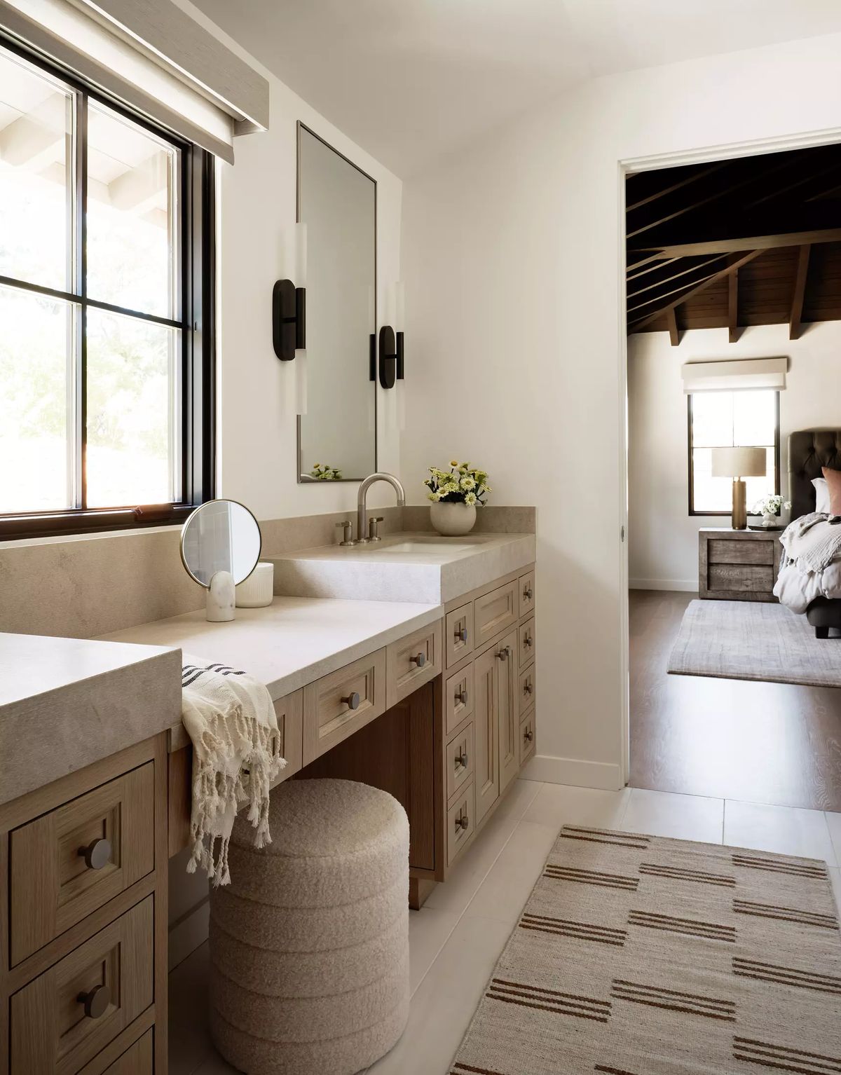 Bathroom with double sink vanity, wooden cabinetry, round mirror, upholstered stool, and light-colored flooring