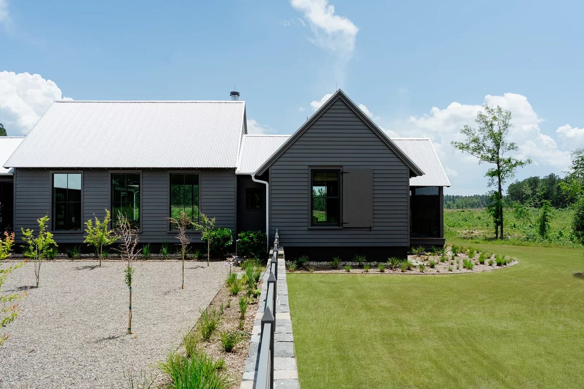 Front exterior view of a gray house with metal roof, multiple windows, gravel pathway, and lawn area.
