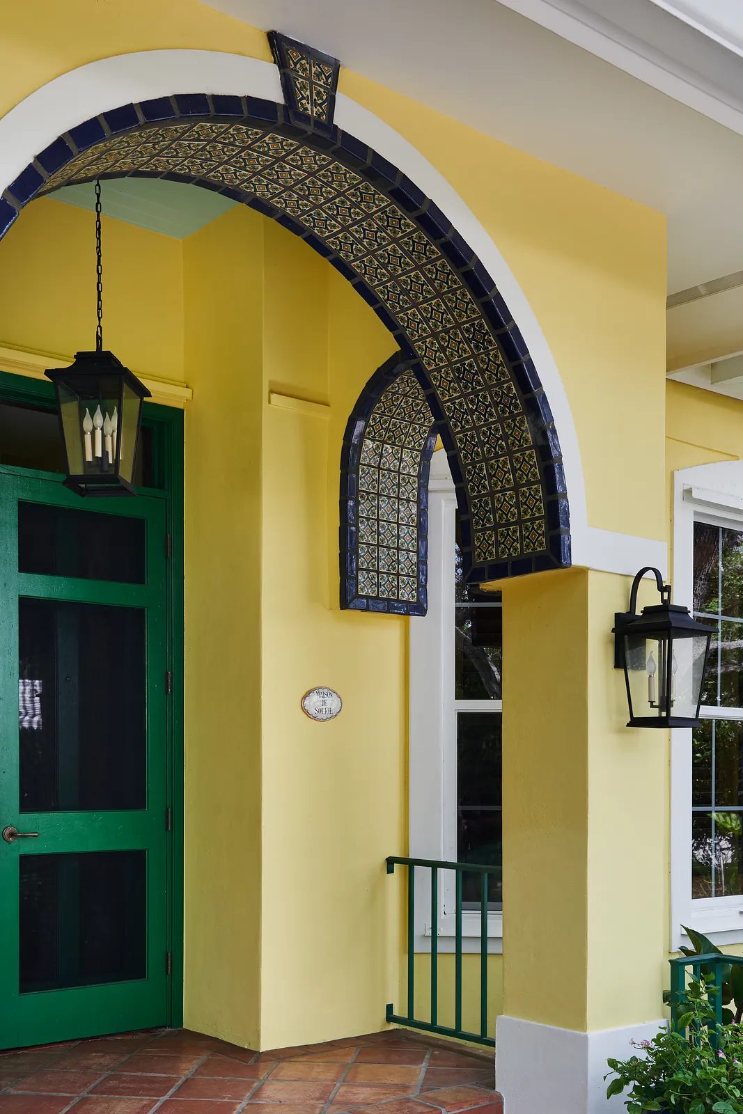Exterior front with yellow wall, blue tiled arches, green doors, large windows, and decorative lanterns