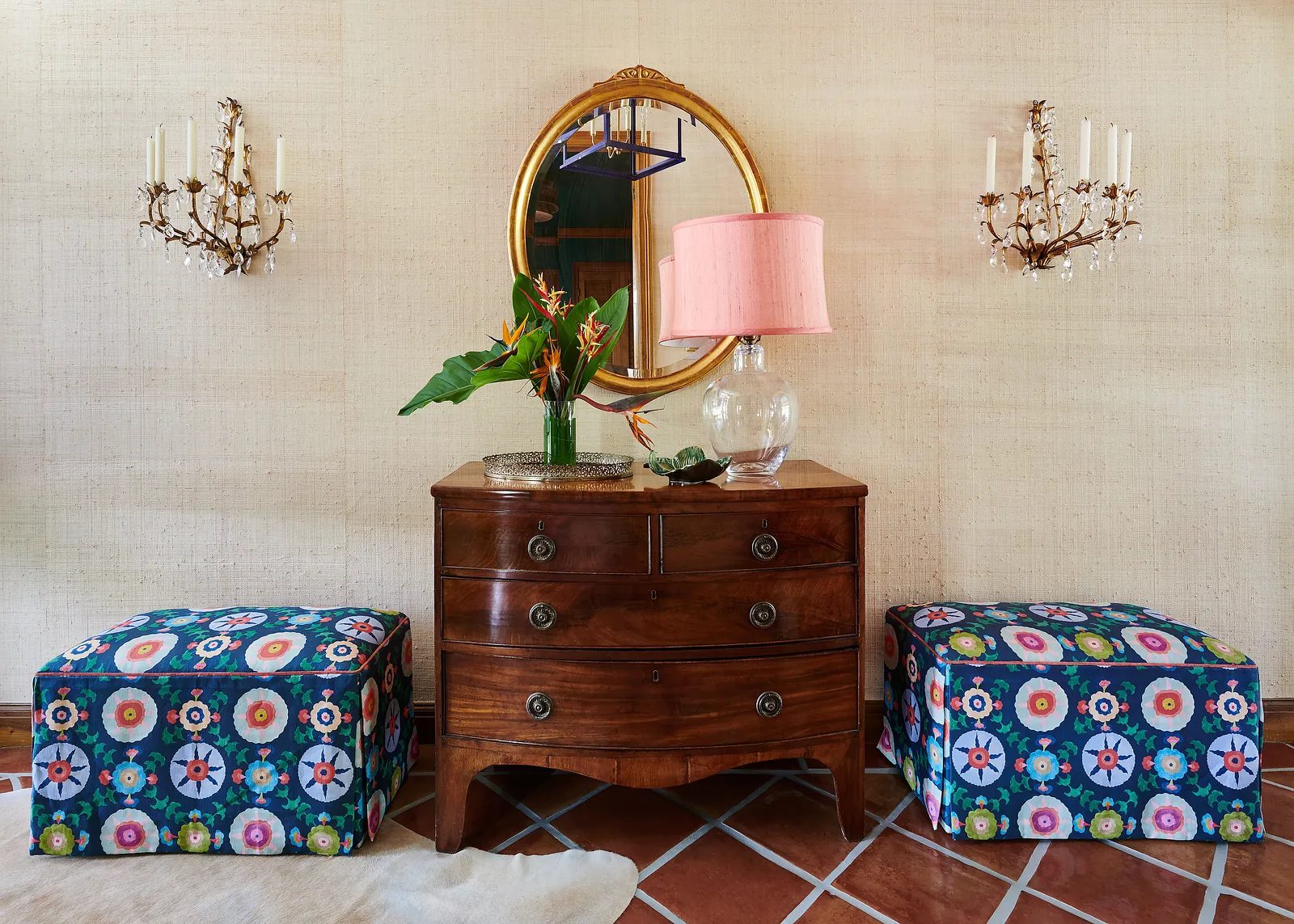 Entryway with wooden chest, patterned ottomans, mirror, wall sconces, and tropical plants.