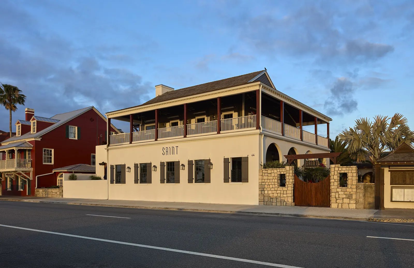 Two-story building with beige facade, black shutters, balcony, arched windows, and stone wall.