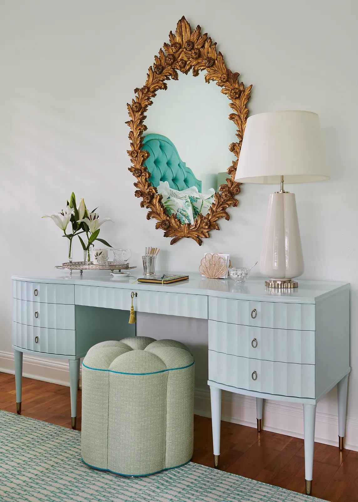 Vanity area with light blue desk, gold mirror, ottoman, lilies in vase, and textured area rug