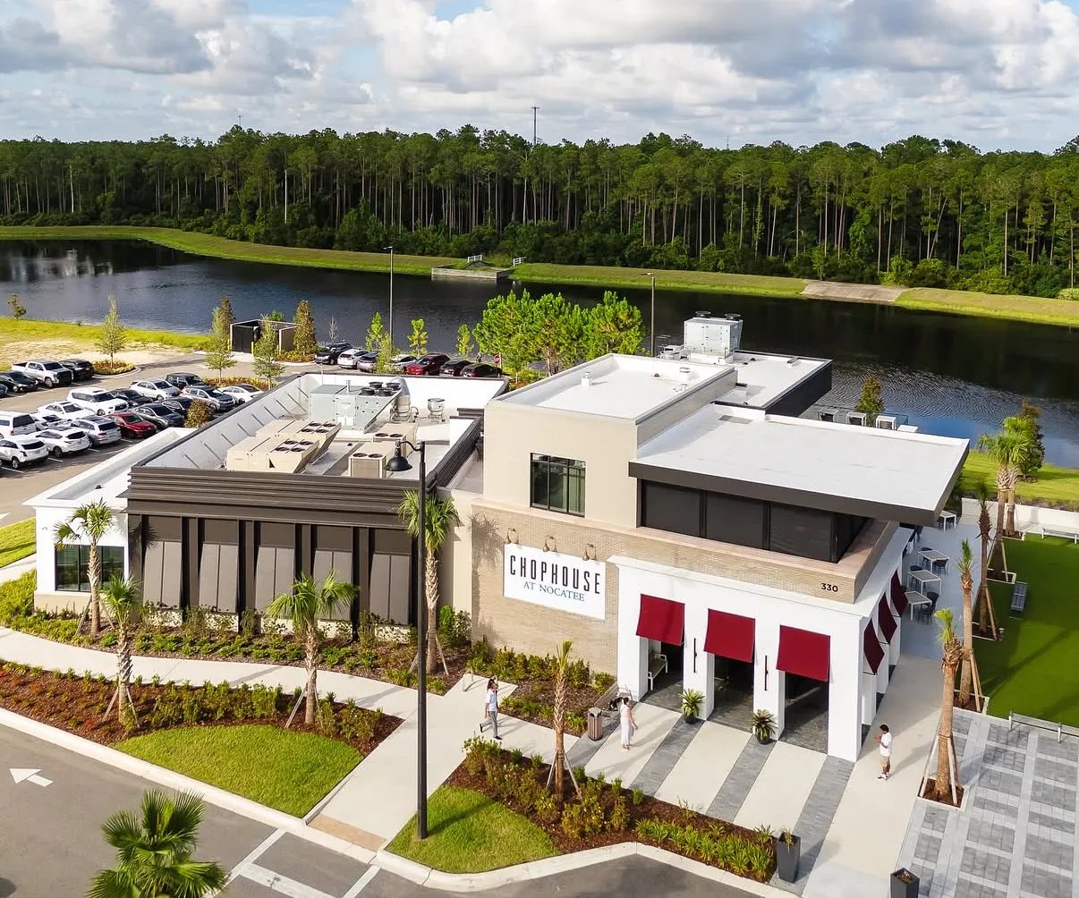 Restaurant building with modern design, large windows, outdoor seating, and manicured landscaping next to a lake.