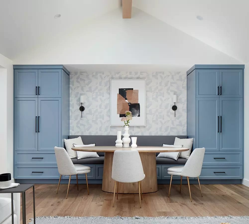 Dining room with circular wooden table, light-colored chairs, blue cabinets, artwork, and wall sconces.