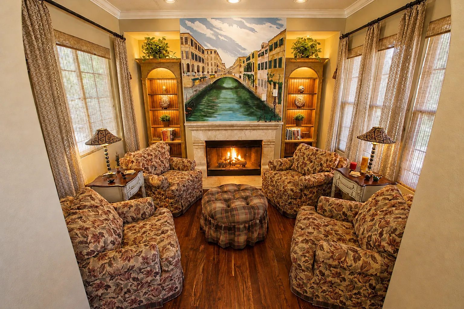 Living room with floral armchairs, central ottoman, fireplace, bookshelves, and a mural of a canal.