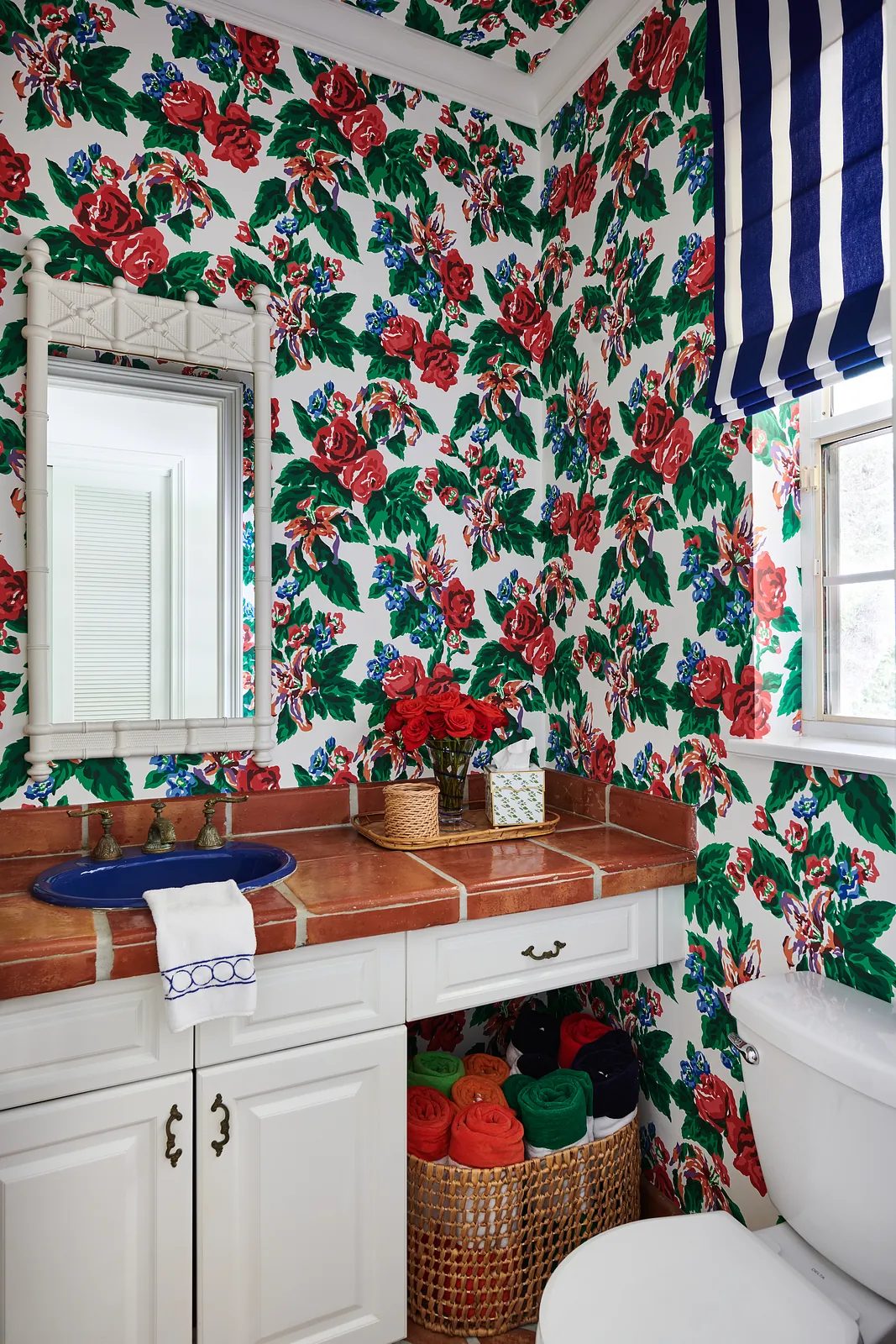 Powder room with floral wallpaper, tiled countertop, mirror, and colorful rolled towels in a basket.