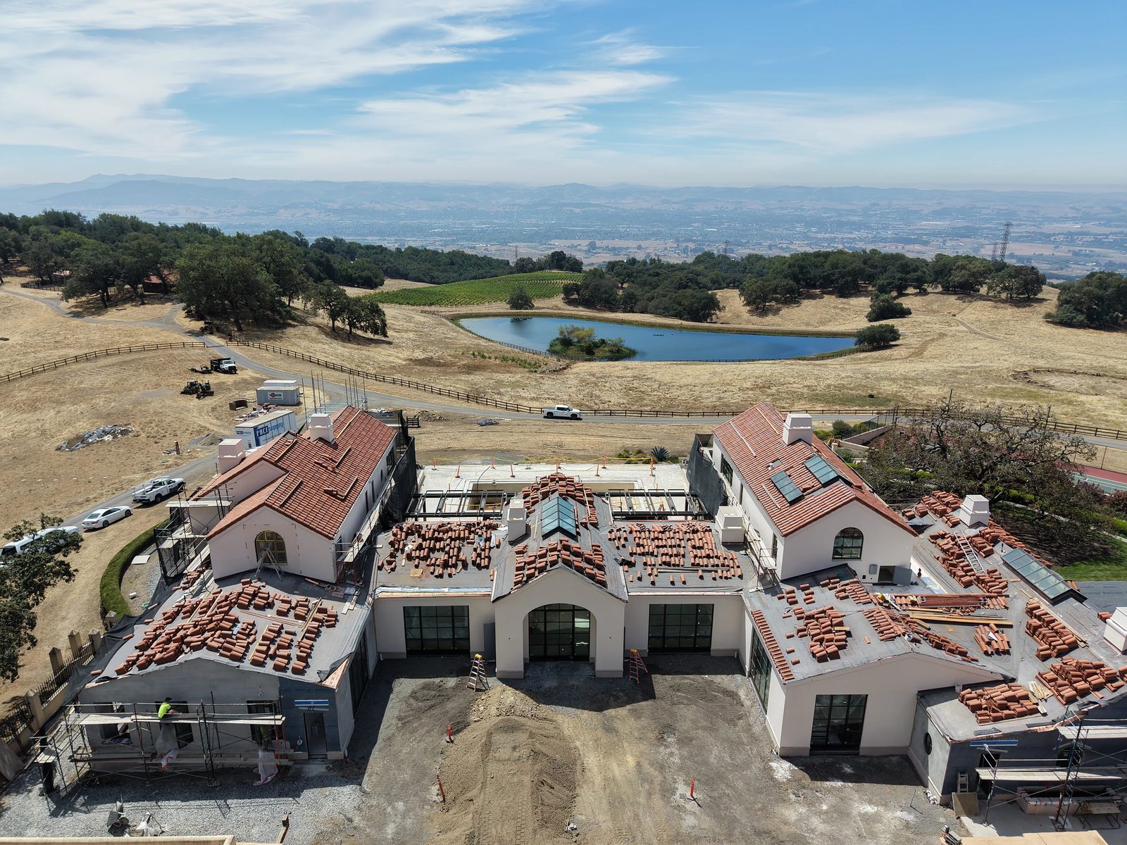 Construction site of a two-story building with tiled roof, windows, and arched doorways near a pond.