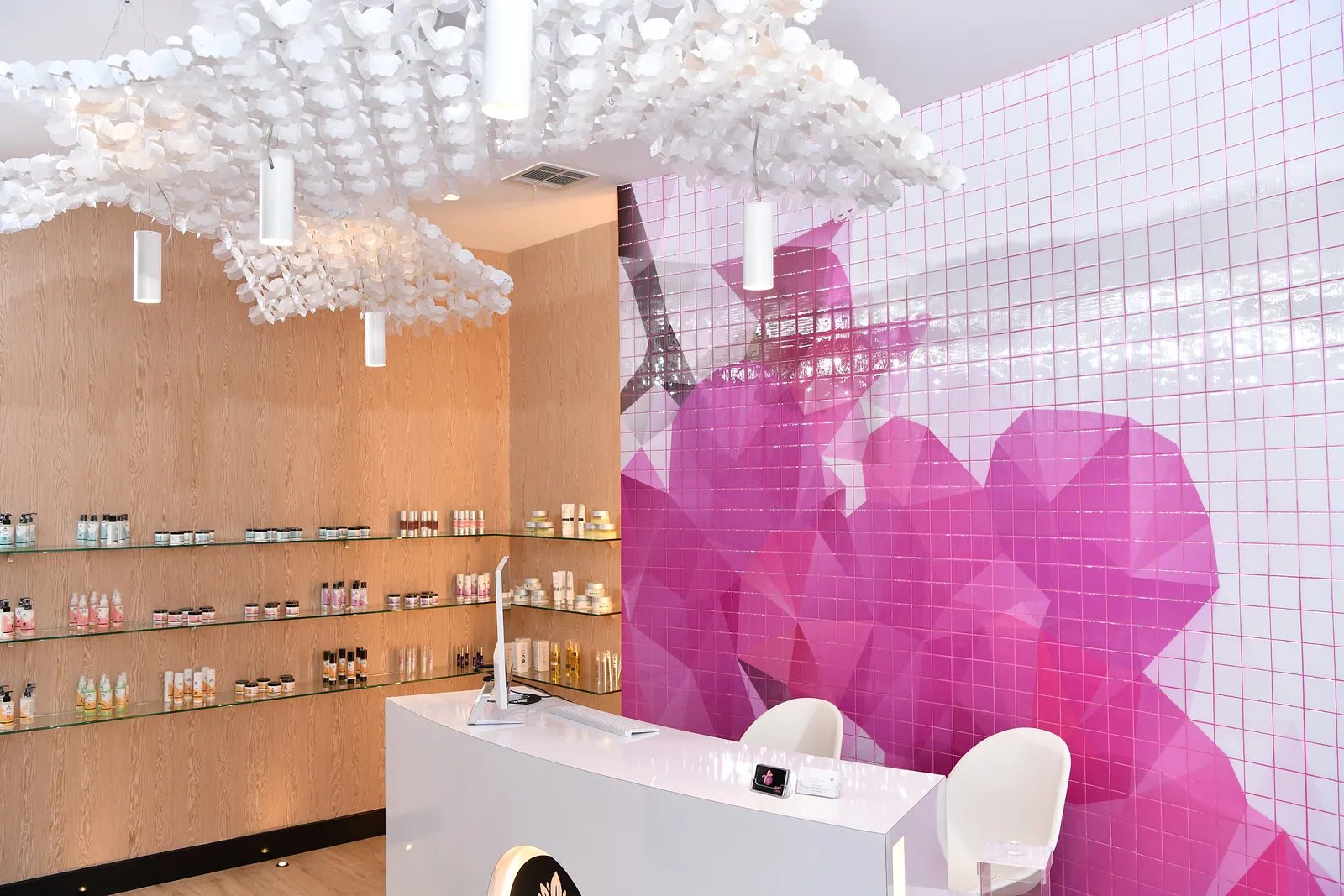 Reception area with white desk, pink mural, shelves of bottles, and cloud-like ceiling installation.