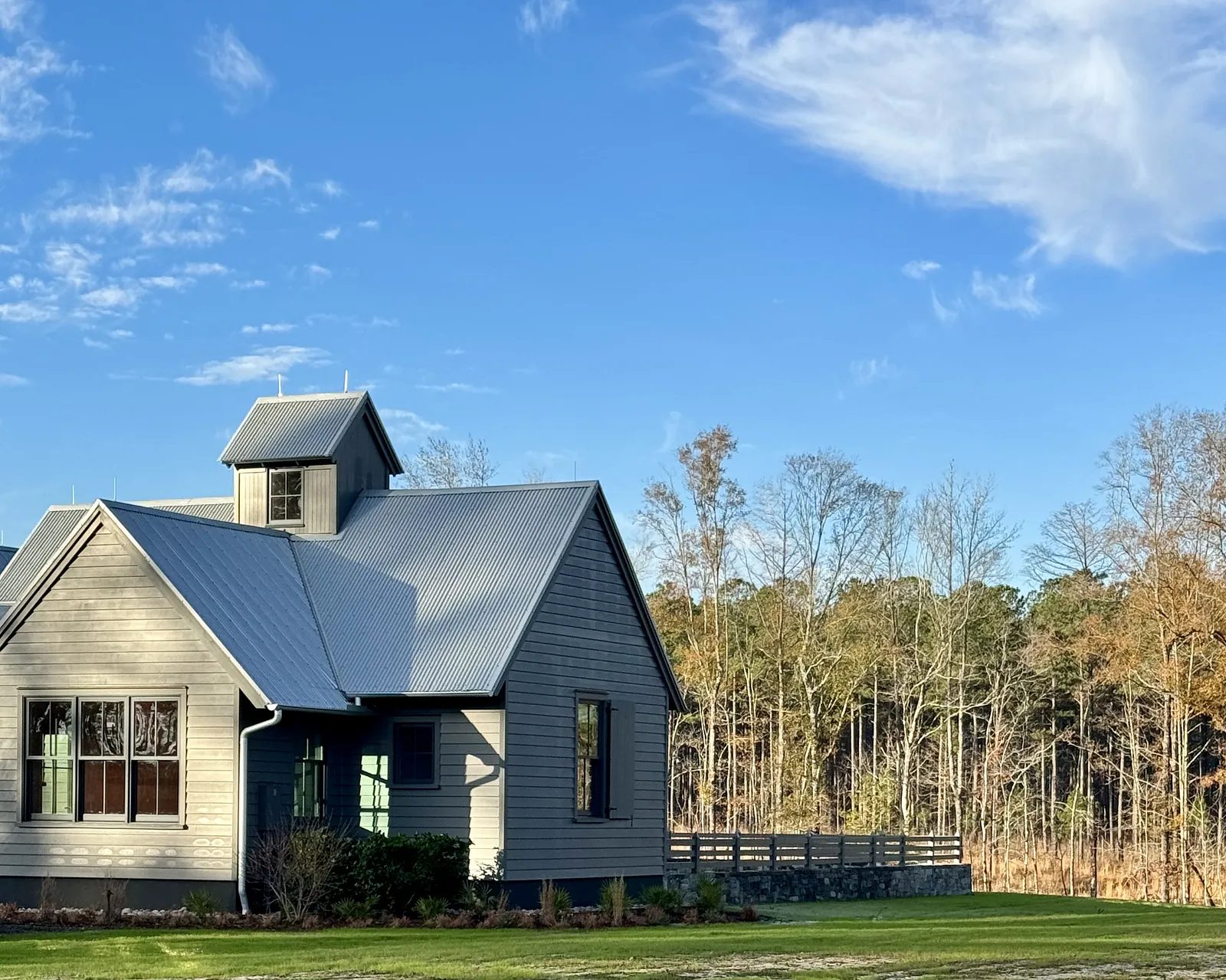 Front exterior of a gray house with gables, metal roof, green windows, grass, and wooden railing.