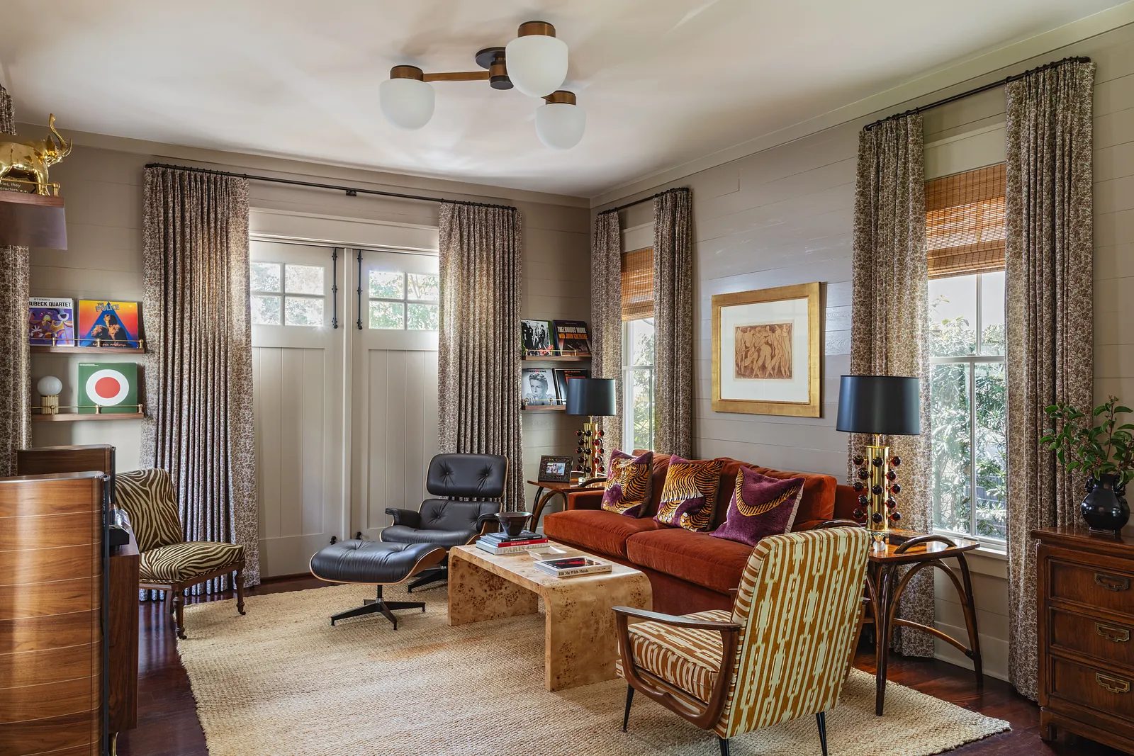 Living room with reddish-brown sofa, patterned armchairs, wood coffee table, artwork shelves, and large windows.
