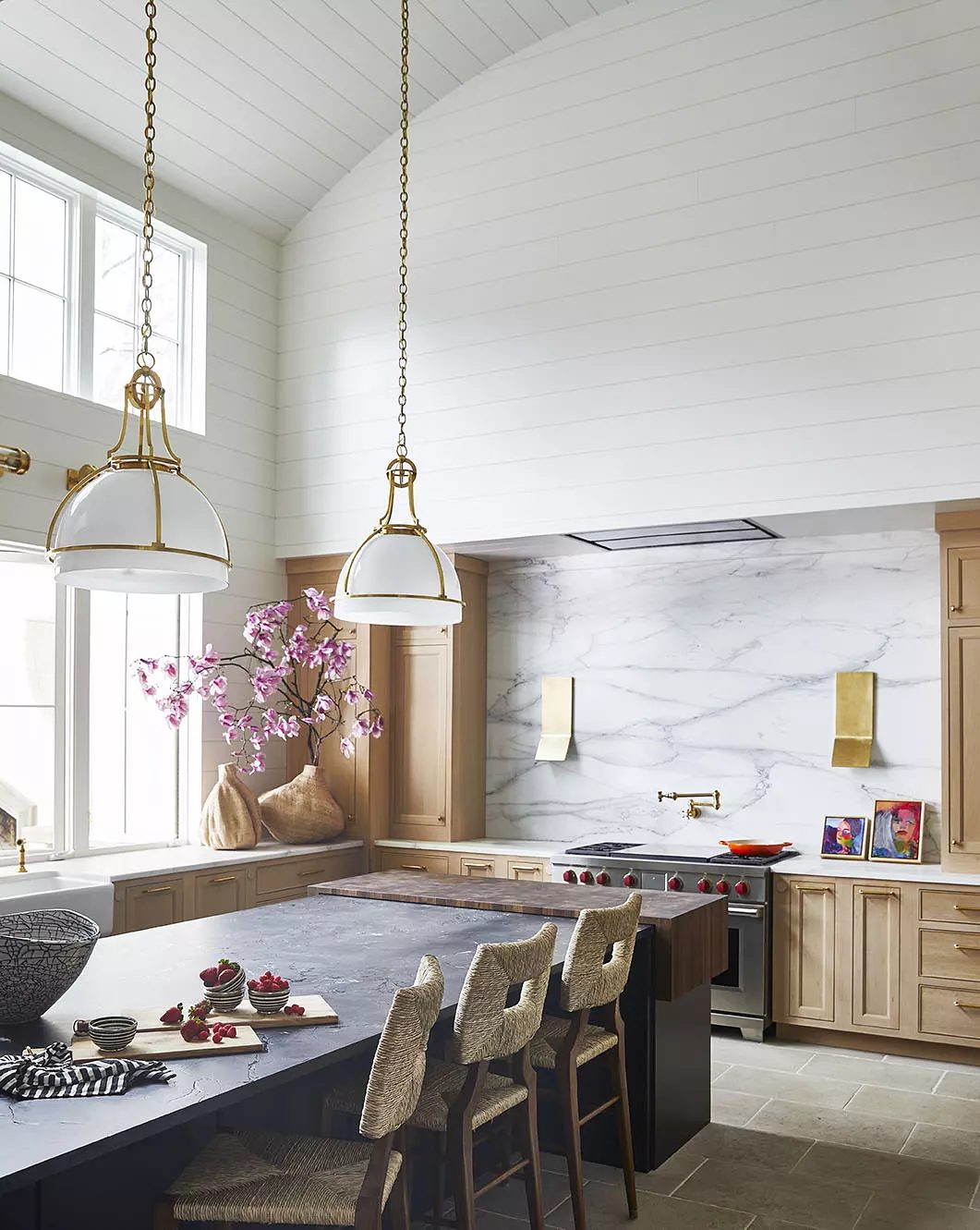 Kitchen with central island, bar stools, light wood cabinetry, marble backsplash, and large windows with flowers.