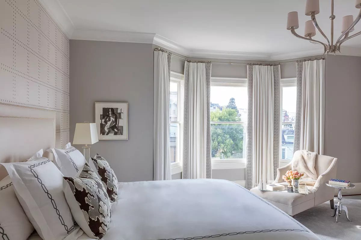 Bedroom with bed, decorative pillows, chair, side table, windows, artwork, and flowers.
