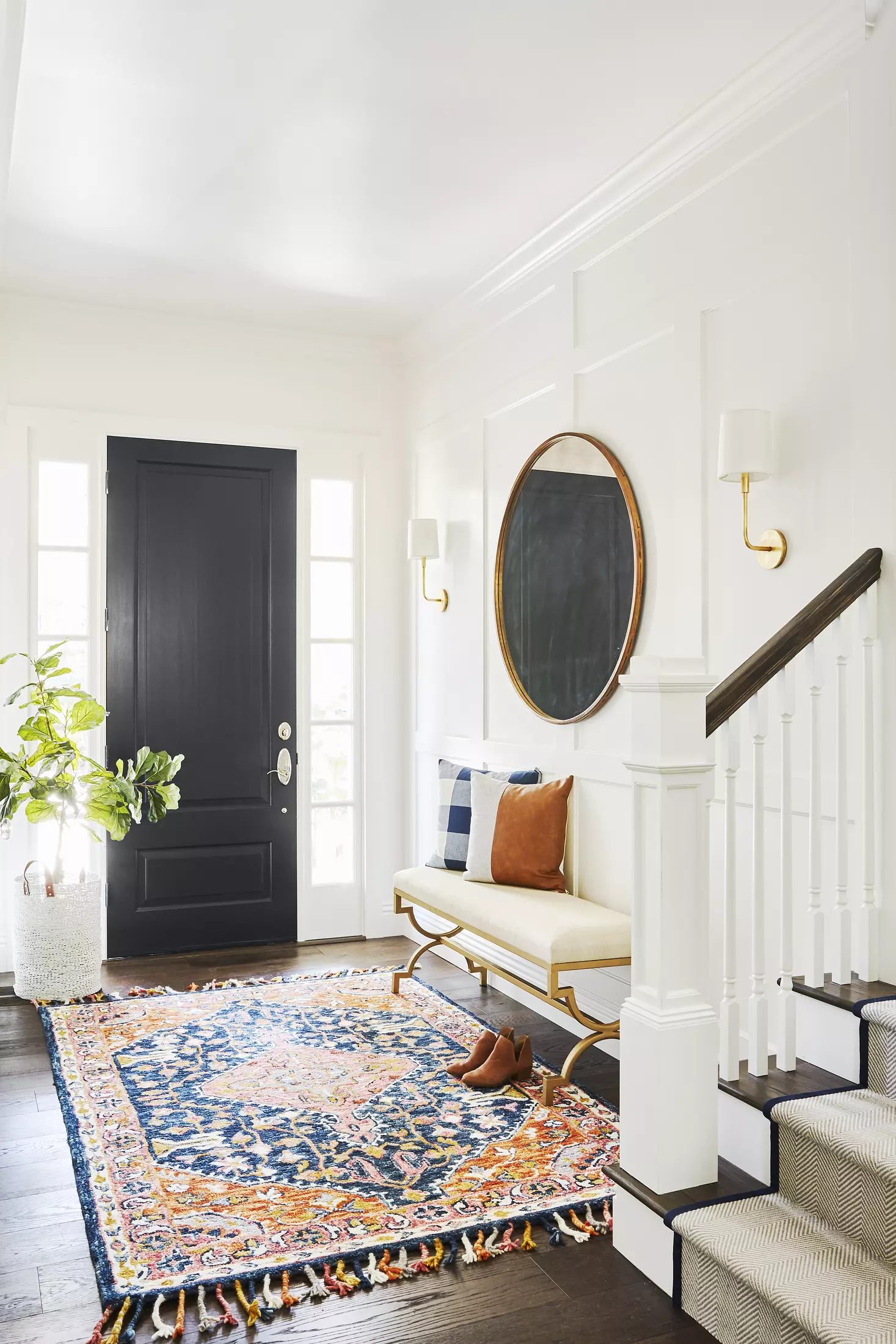 Foyer with black door, white sofa, round mirror, wall sconces, staircase, and colorful area rug