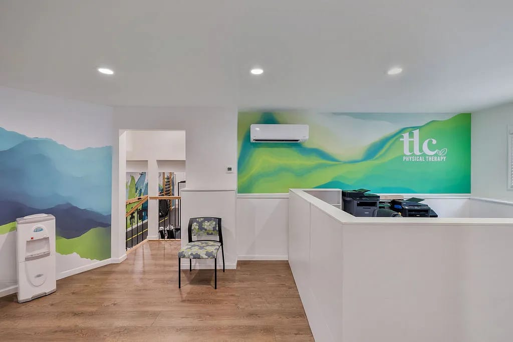 Office reception area with white desk, patterned chair, colorful wall murals, and water cooler.