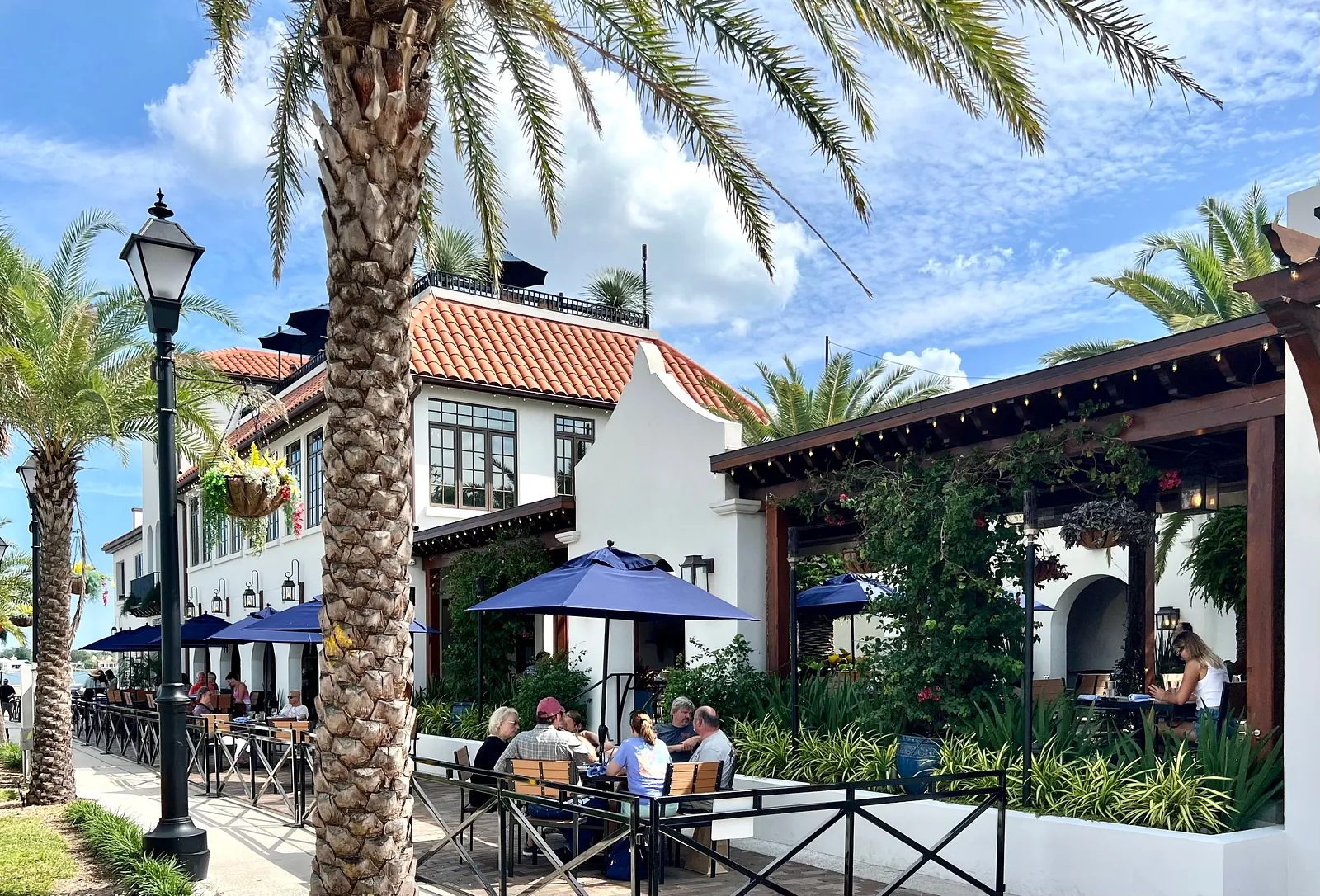 Outdoor patio with tables, chairs, blue umbrellas, palm trees, and a building with clay tile roof.