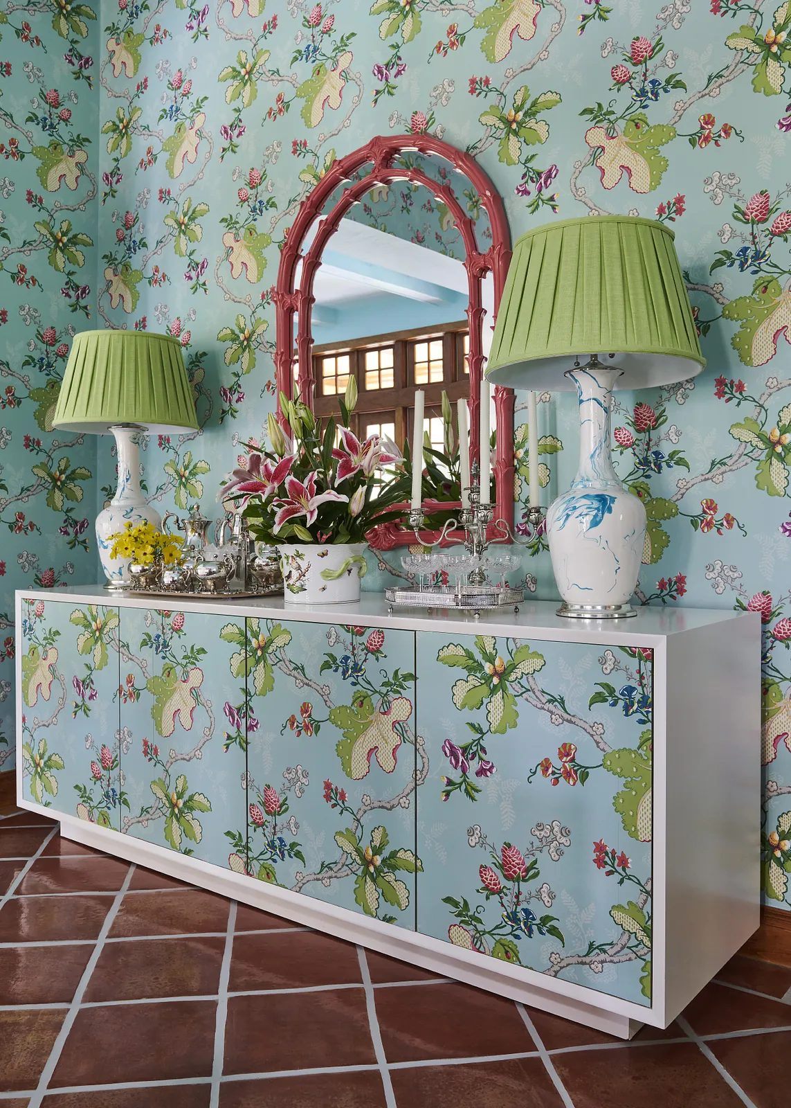 Foyer with white sideboard, floral wallpaper, green lamps, round mirror, and patterned tile floor.
