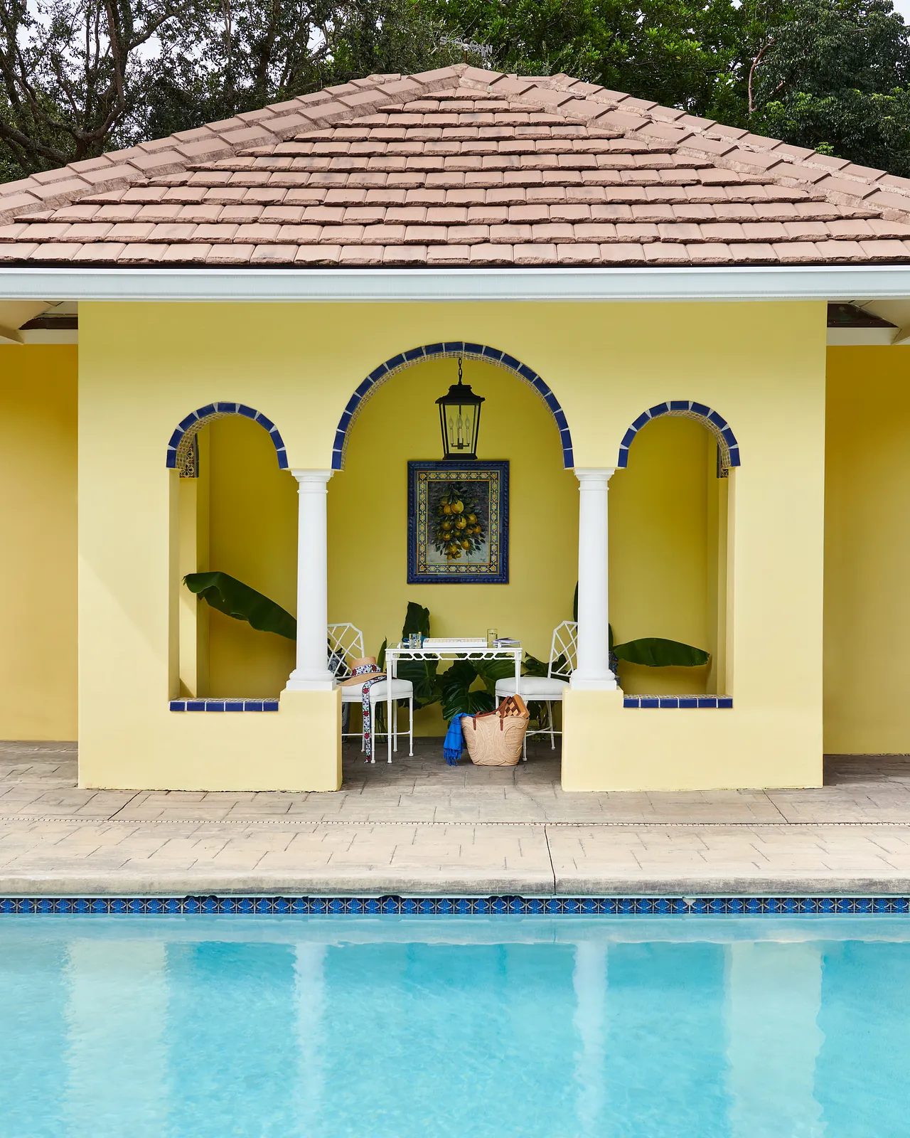 Patio with yellow walls, arched openings, light wood furniture, framed artwork, and a swimming pool.