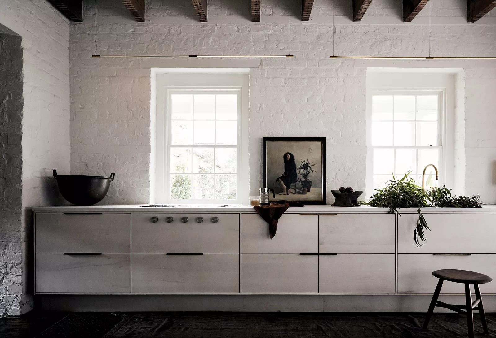 Kitchen with light cabinetry, windows, artwork, herbs, and a stool in front.