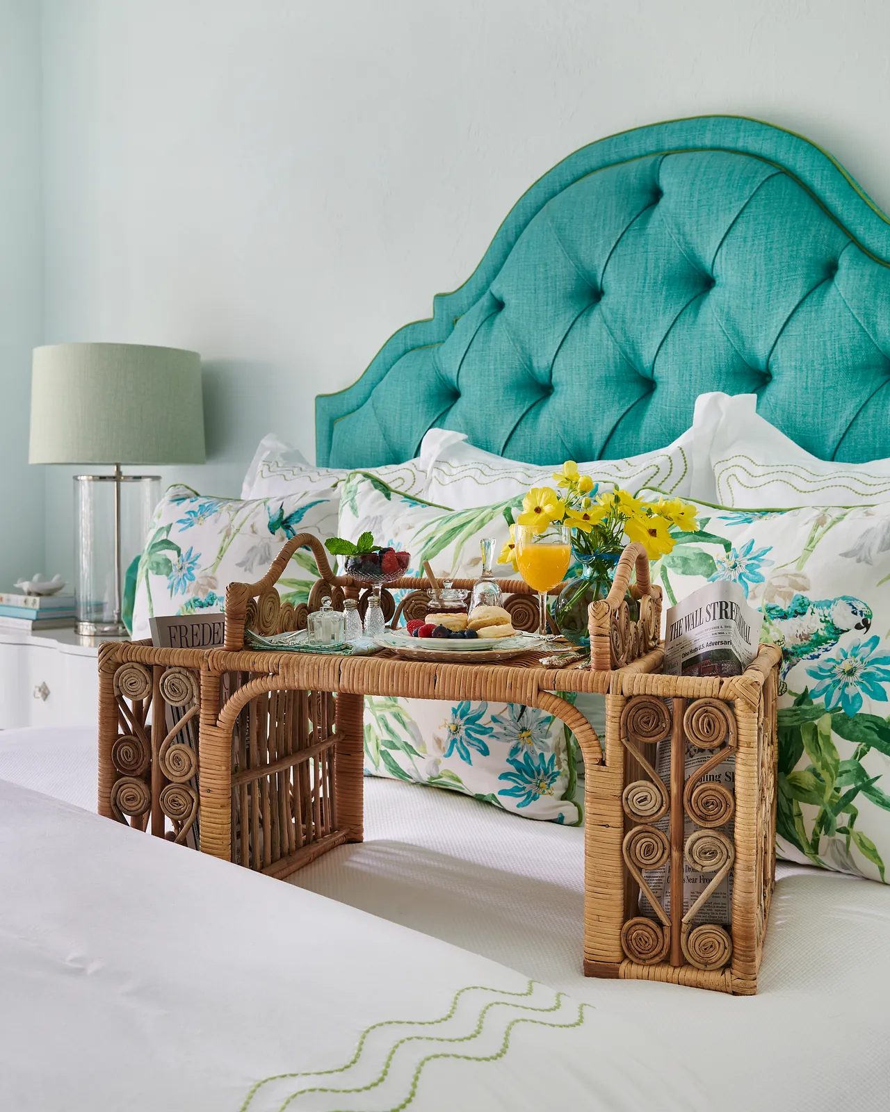 Bedroom with turquoise headboard, white bedding, wicker tray, and bedside table with green lamp.