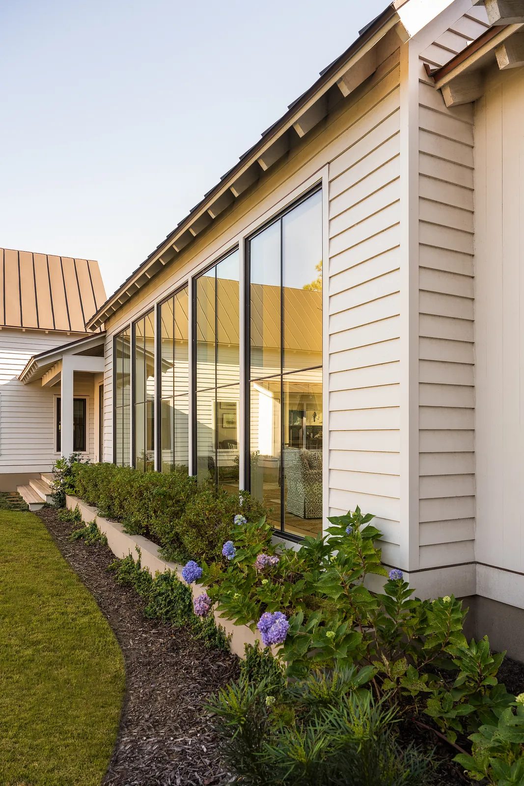 Exterior rear view of a house with large windows, white siding, brown metal roof, and landscaped garden.
