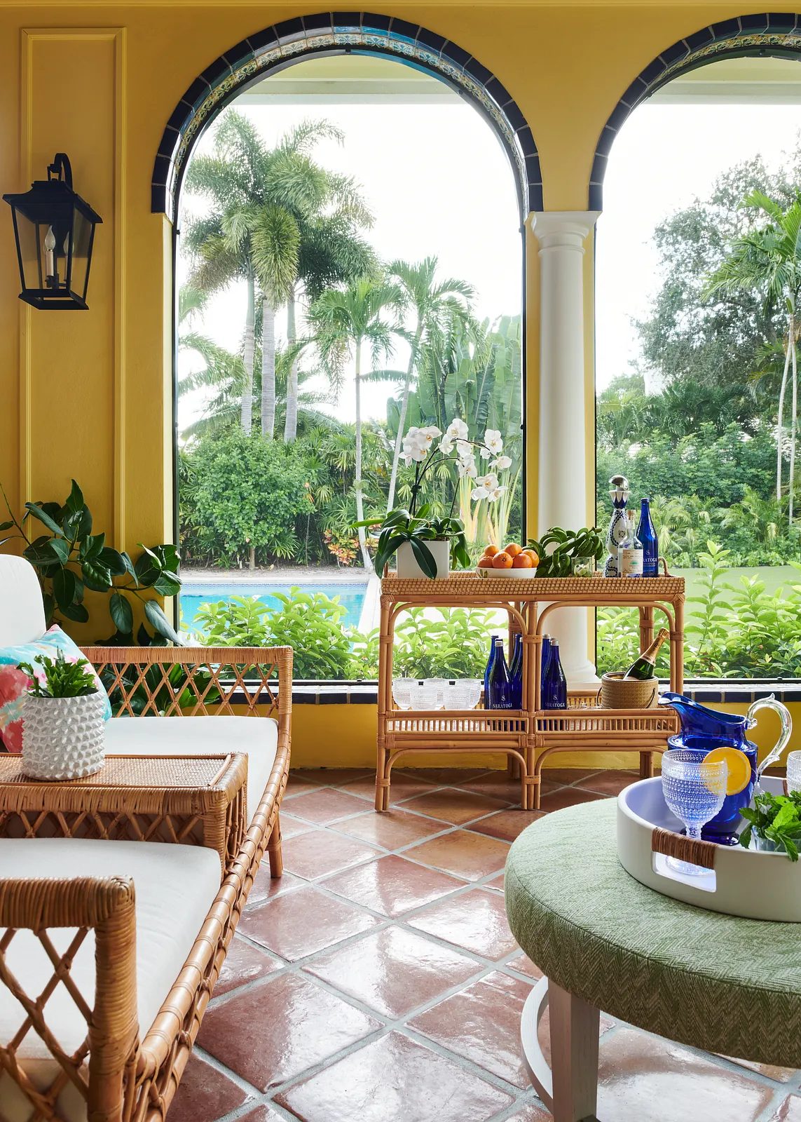 Sunroom with rattan furniture, potted plants, and large arched windows overlooking greenery.