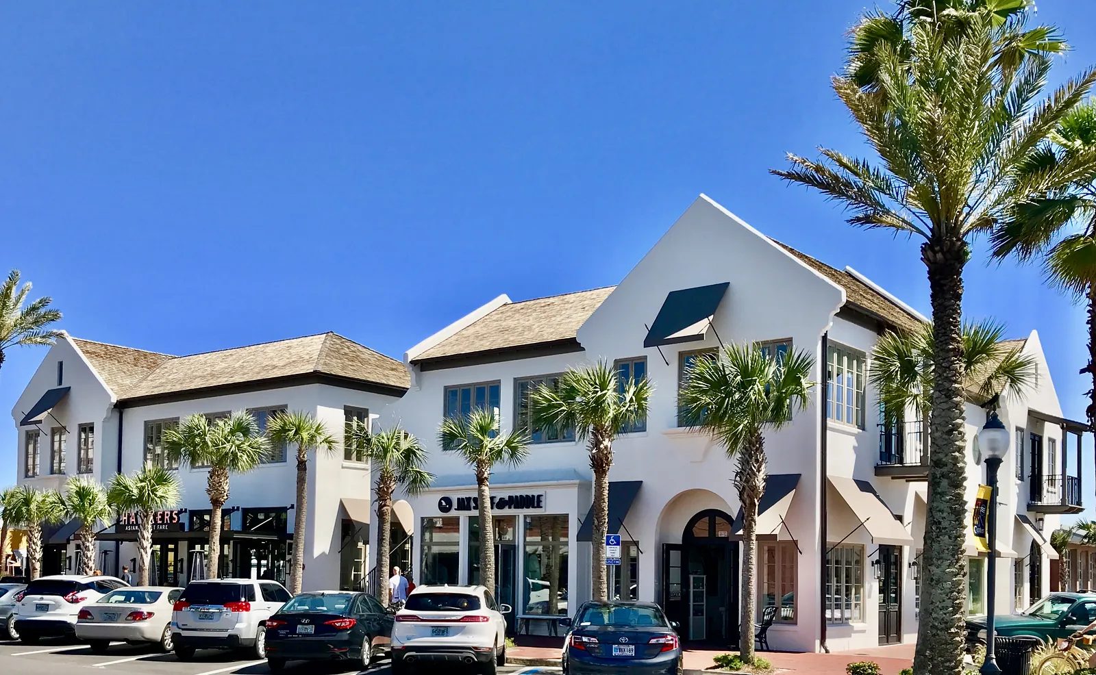 Exterior view of a commercial building with white facade, large windows, palm trees, and parked cars.
