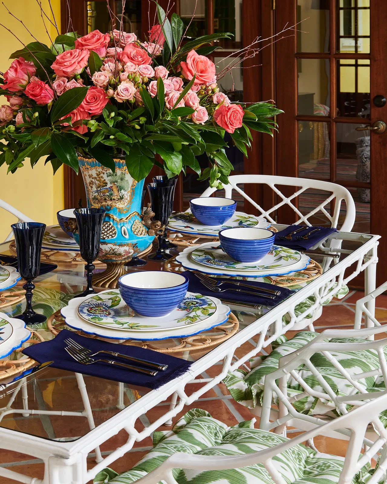 Dining room with glass-top table, wicker chairs, blue dinnerware, black glassware, and floral centerpiece.