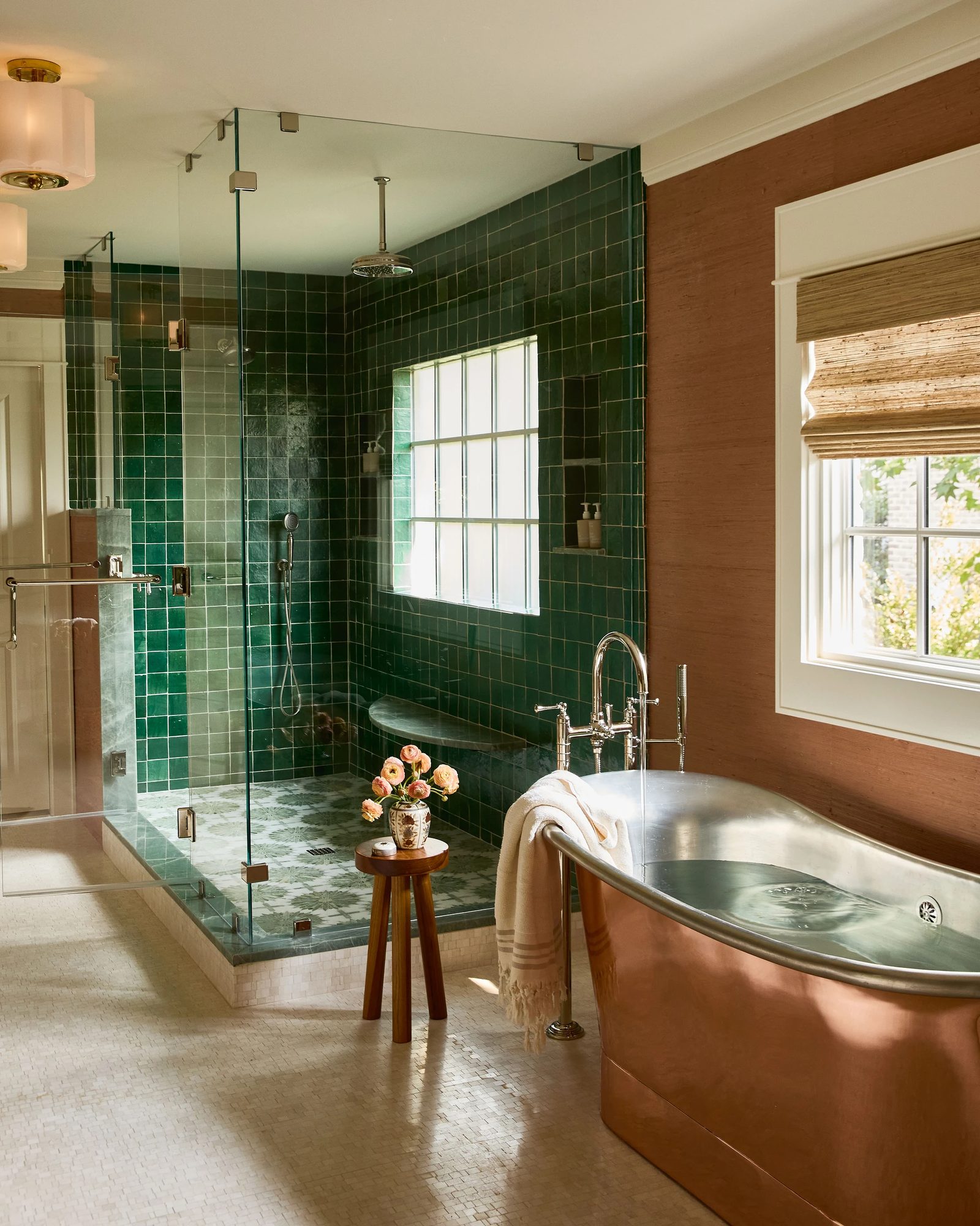 Bathroom with copper bathtub, glass shower, green tile, wooden stool with flowers, and window with woven blind.