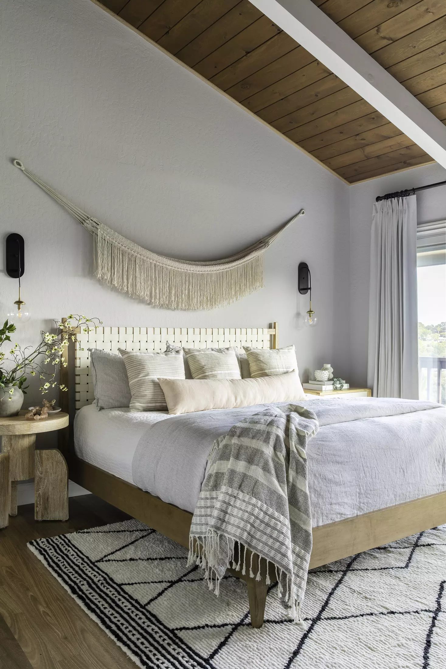 Bedroom with wooden bed, white bedspread, decorative pillows, side table, and geometric area rug with natural light.