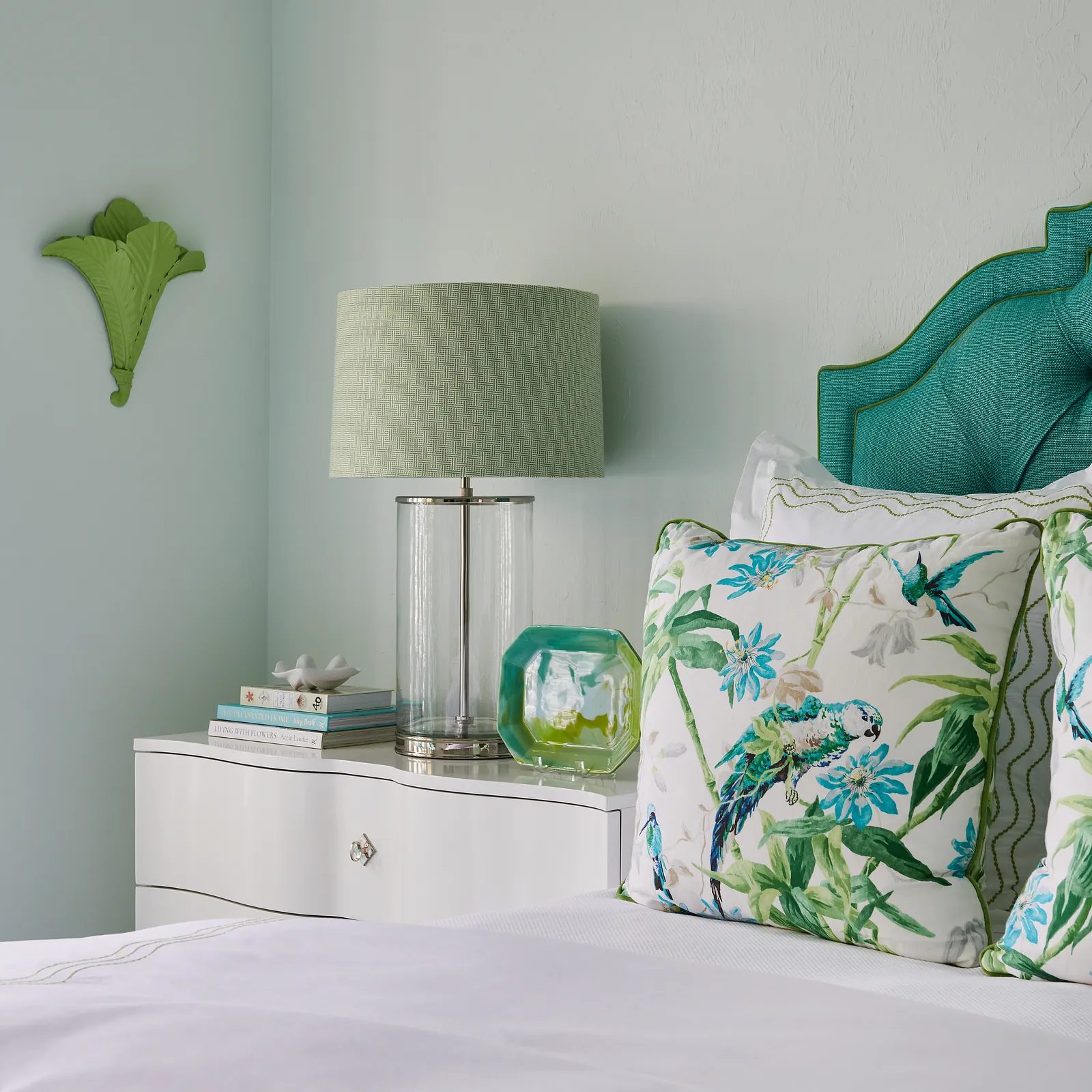 Bedroom with floral headboard, nightstand, lamp, and decorative pillows on a light green wall.