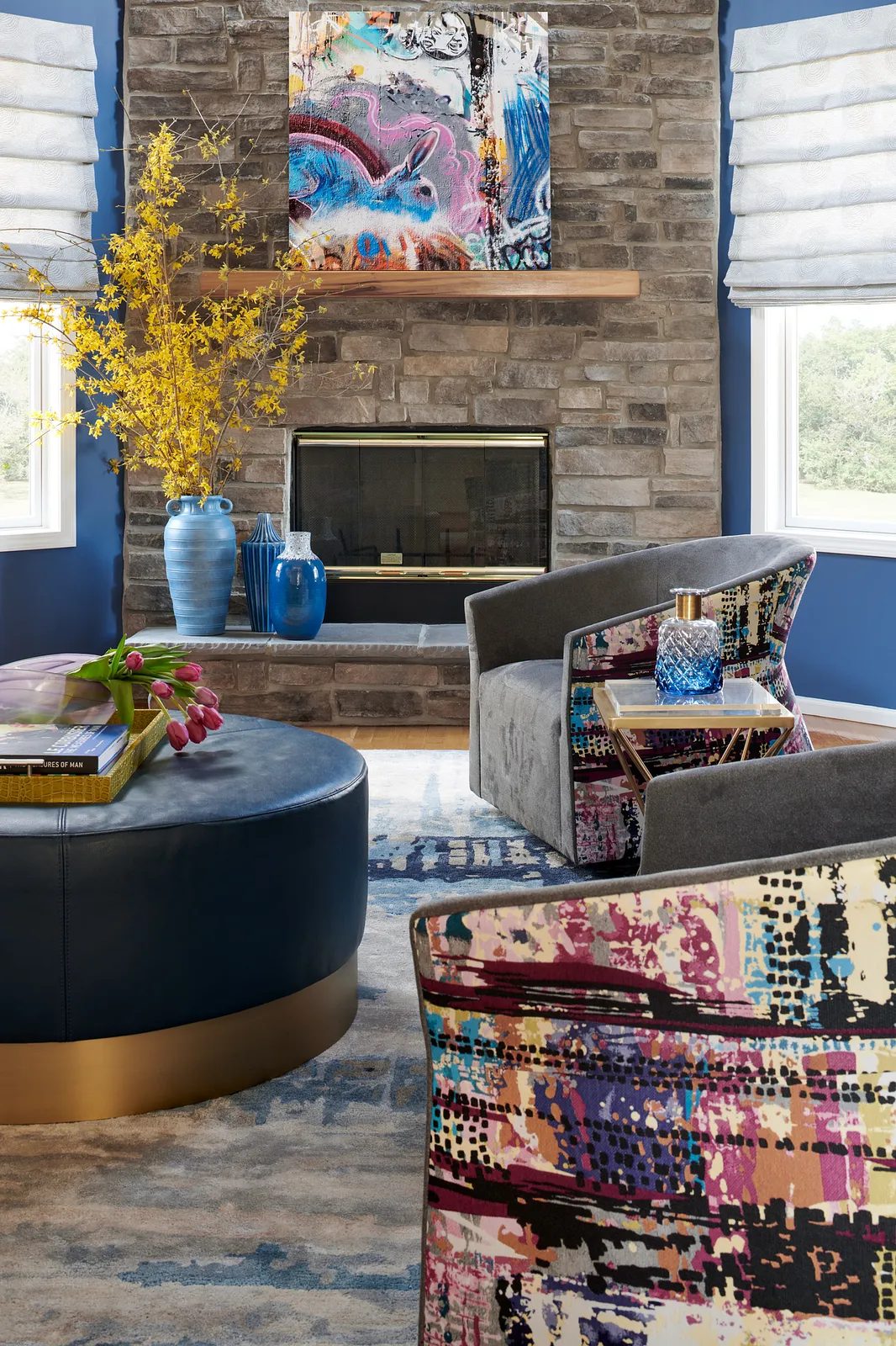 Living room with round ottoman, modern chairs, stone fireplace, blue walls, and yellow flower vase.