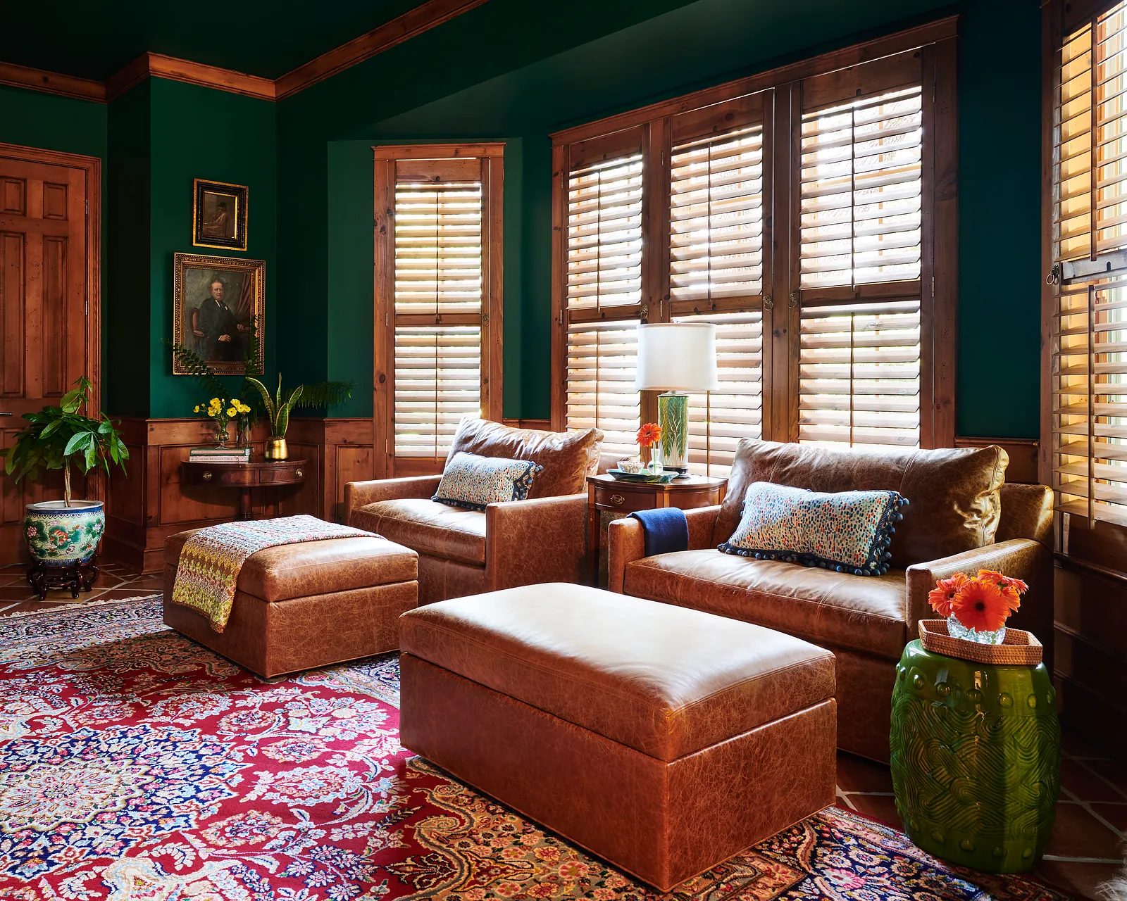 Living room with brown leather armchairs, ottoman, vibrant area rug, and wooden windows with blinds.