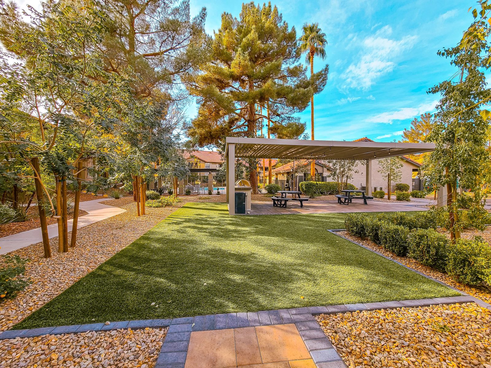 Garden area with artificial turf, covered pavilion, picnic tables, stone pathways, and surrounding greenery