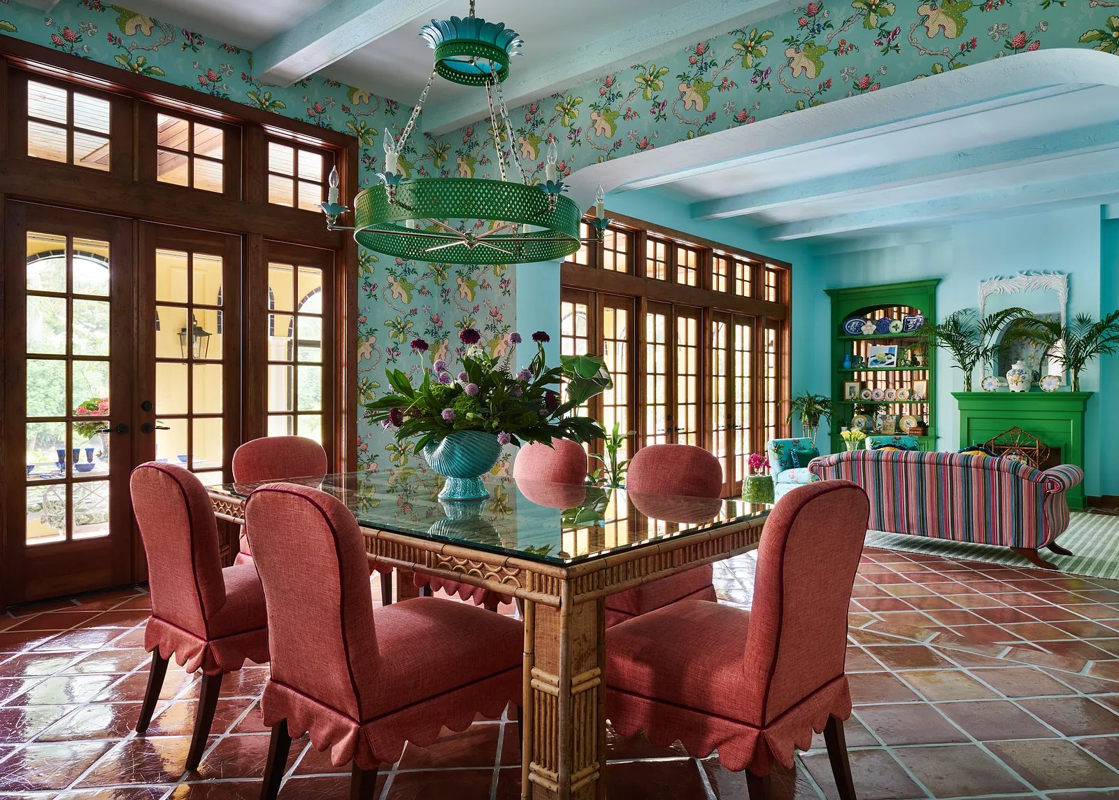 Dining room with glass table, coral chairs, floral wallpaper, green chandelier, and striped sofa.