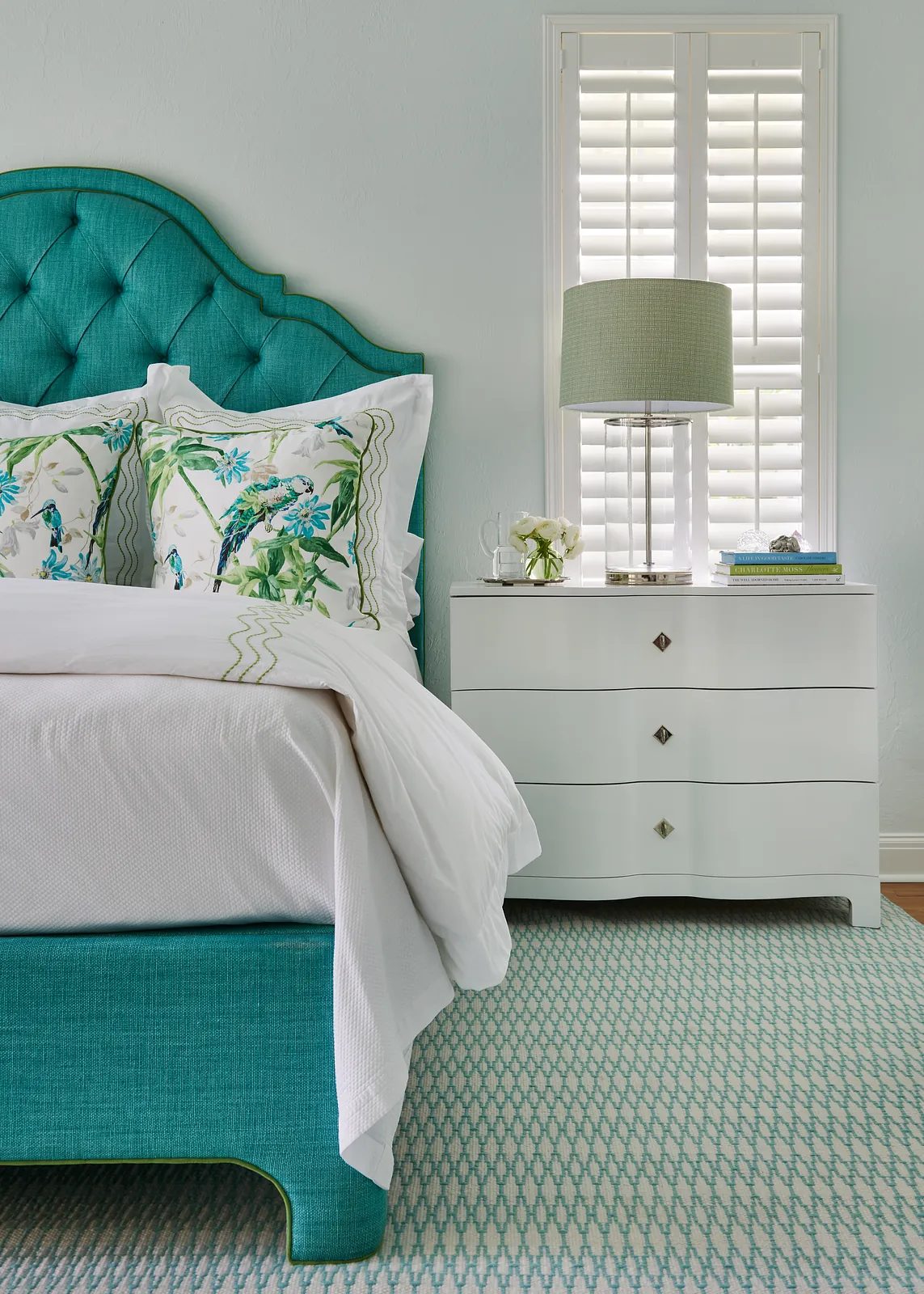 Bedroom with turquoise bed, white nightstand, green lamp, and patterned area rug.