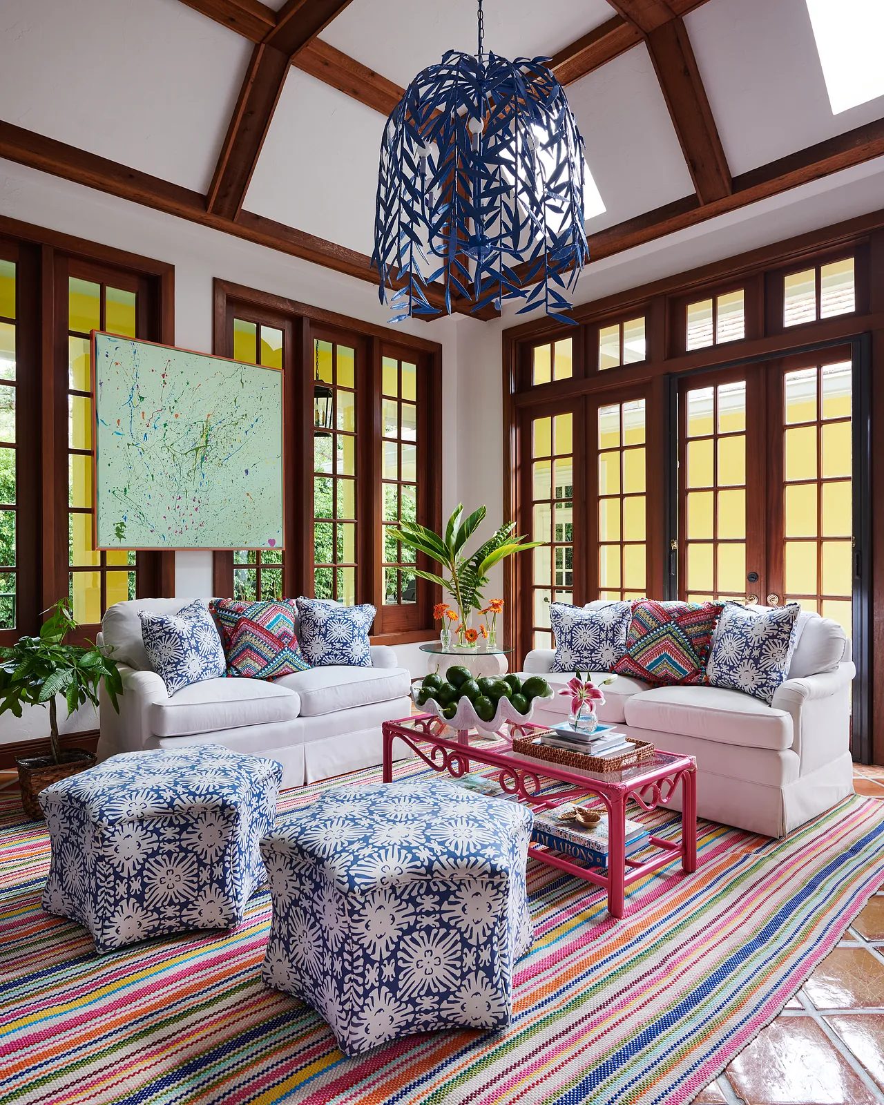 Living room with white sofas, red coffee table, patterned ottomans, large windows, and colorful area rug.