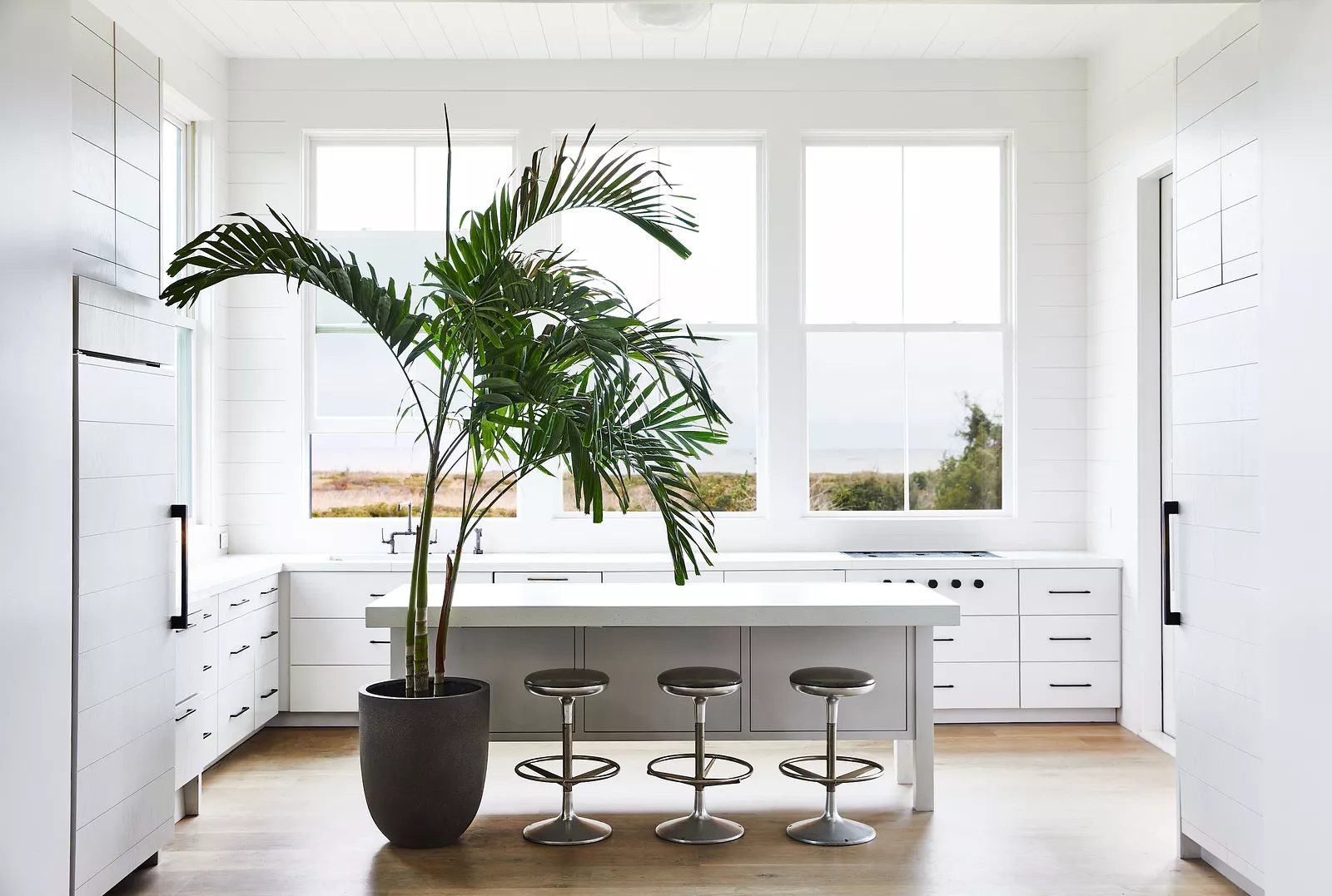 Kitchen with white island, three bar stools, large windows, and a potted plant.