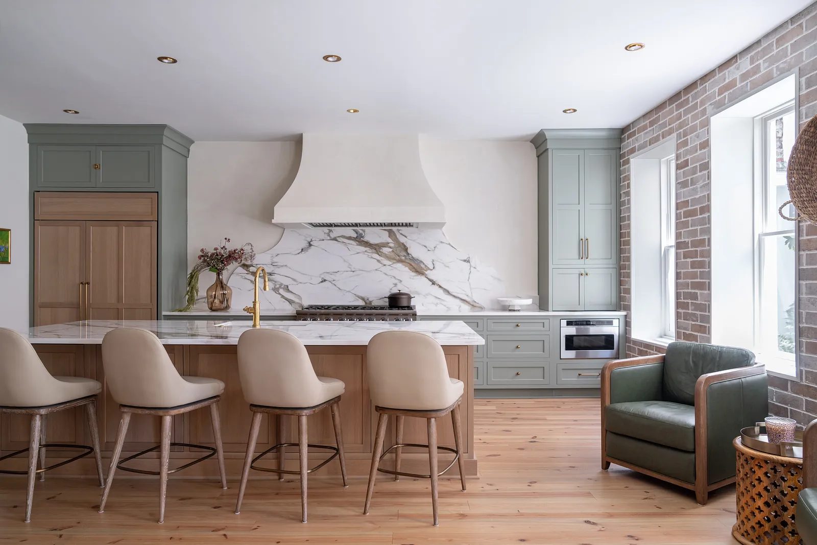 Kitchen with marble island, beige bar stools, green cabinetry, range hood, and large windows.