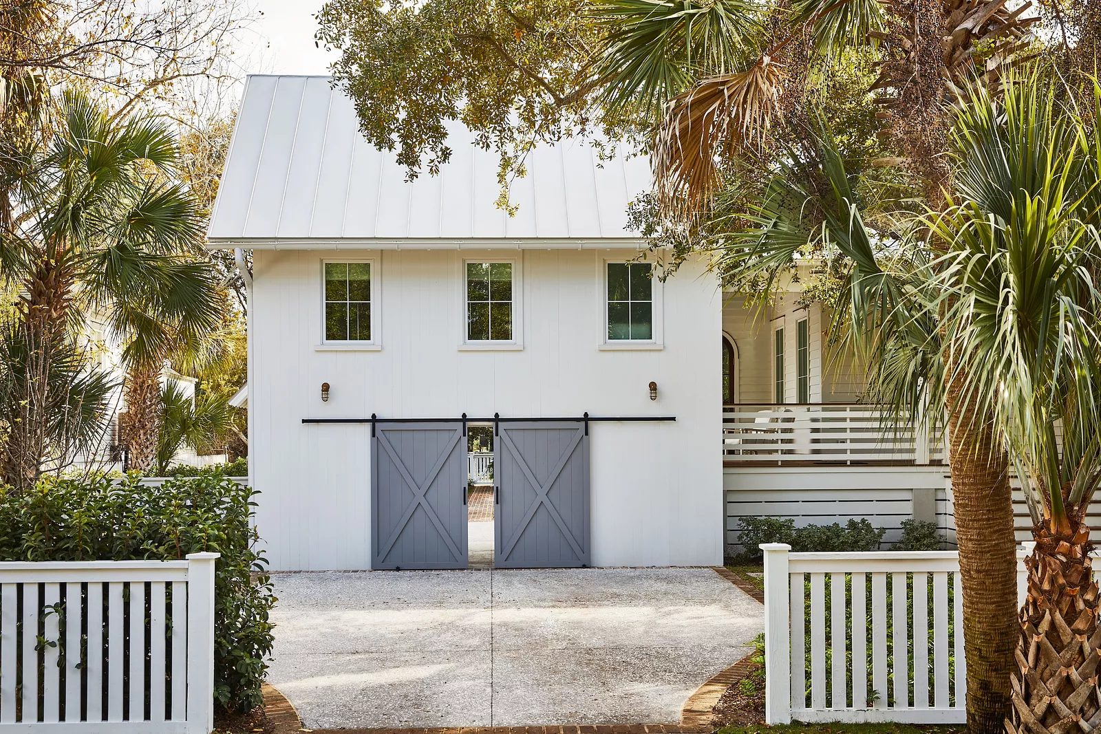 Exterior front view of a white barn-style building with sliding doors, palm trees, and a picket fence.
