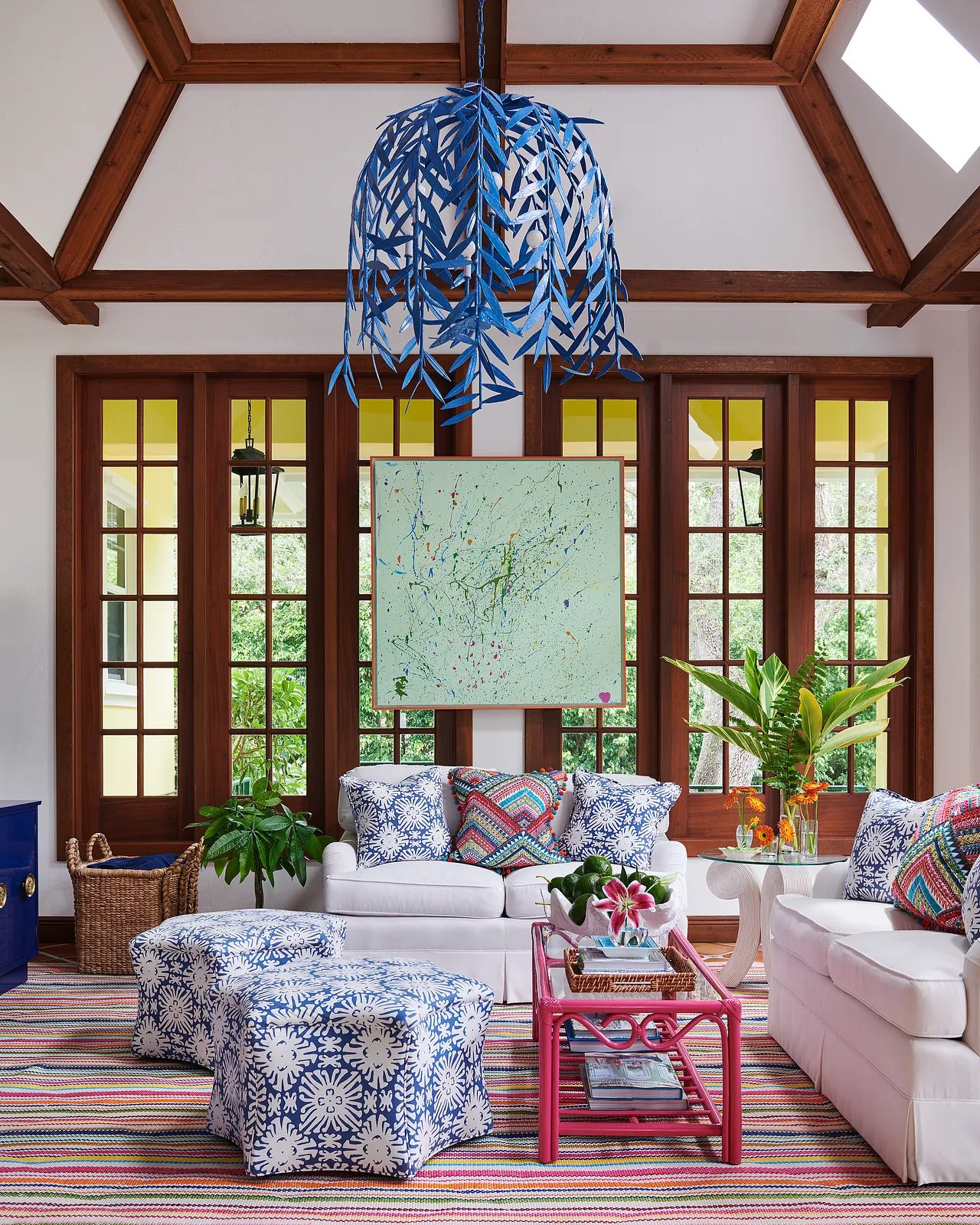 Living room with white sofas, colorful rug, pink coffee table, blue-patterned ottomans, and large windows.
