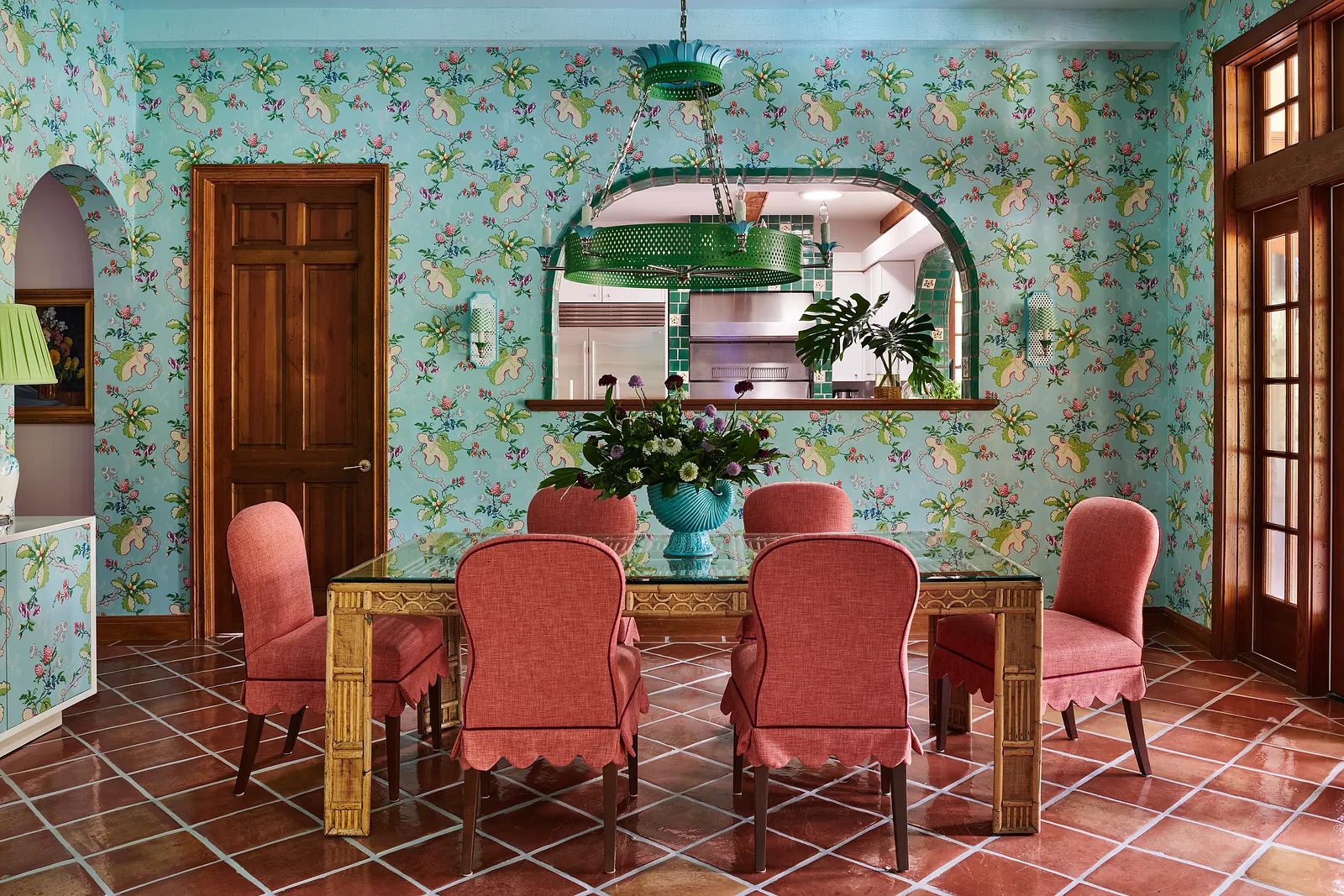 Dining room with glass table, pink chairs, floral wallpaper, arched opening, and terracotta tile floor