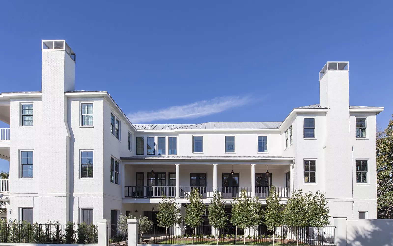 Two-story white house with metal roof, columns, porch, double doors, and small trees in the front garden.
