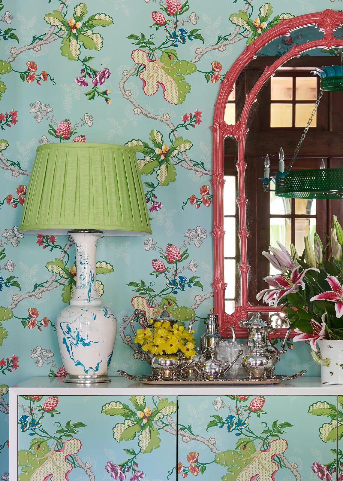 Colorful space with floral wallpaper, white cabinet, green lamp, silver tray, and pink-framed mirror.
