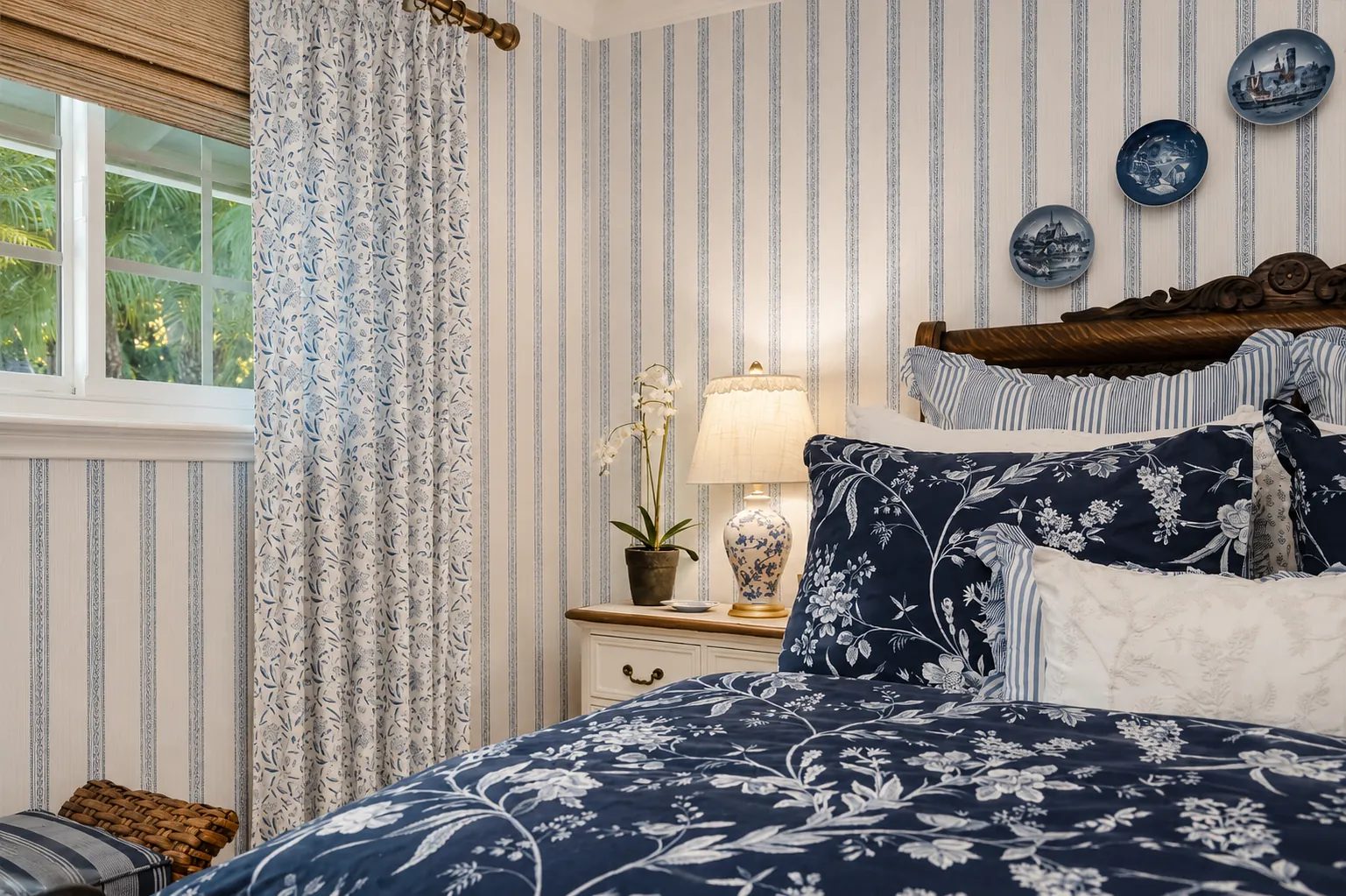 Bedroom with floral-patterned quilt, decorative pillows, nightstand, lamp, and striped walls.
