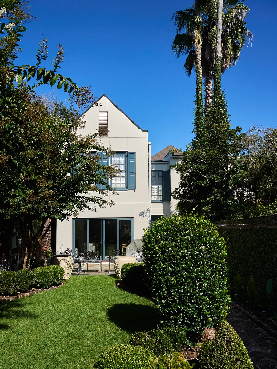 Backyard with grass, round shrubs, palm trees, modern house with blue shutters and large windows.