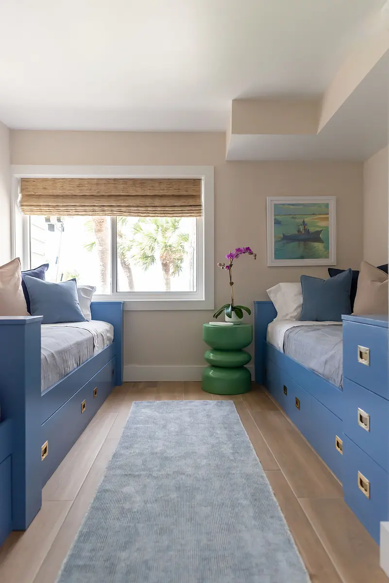 Bedroom with two blue built-in beds, green side table, light blue rug, and large window with bamboo shade.