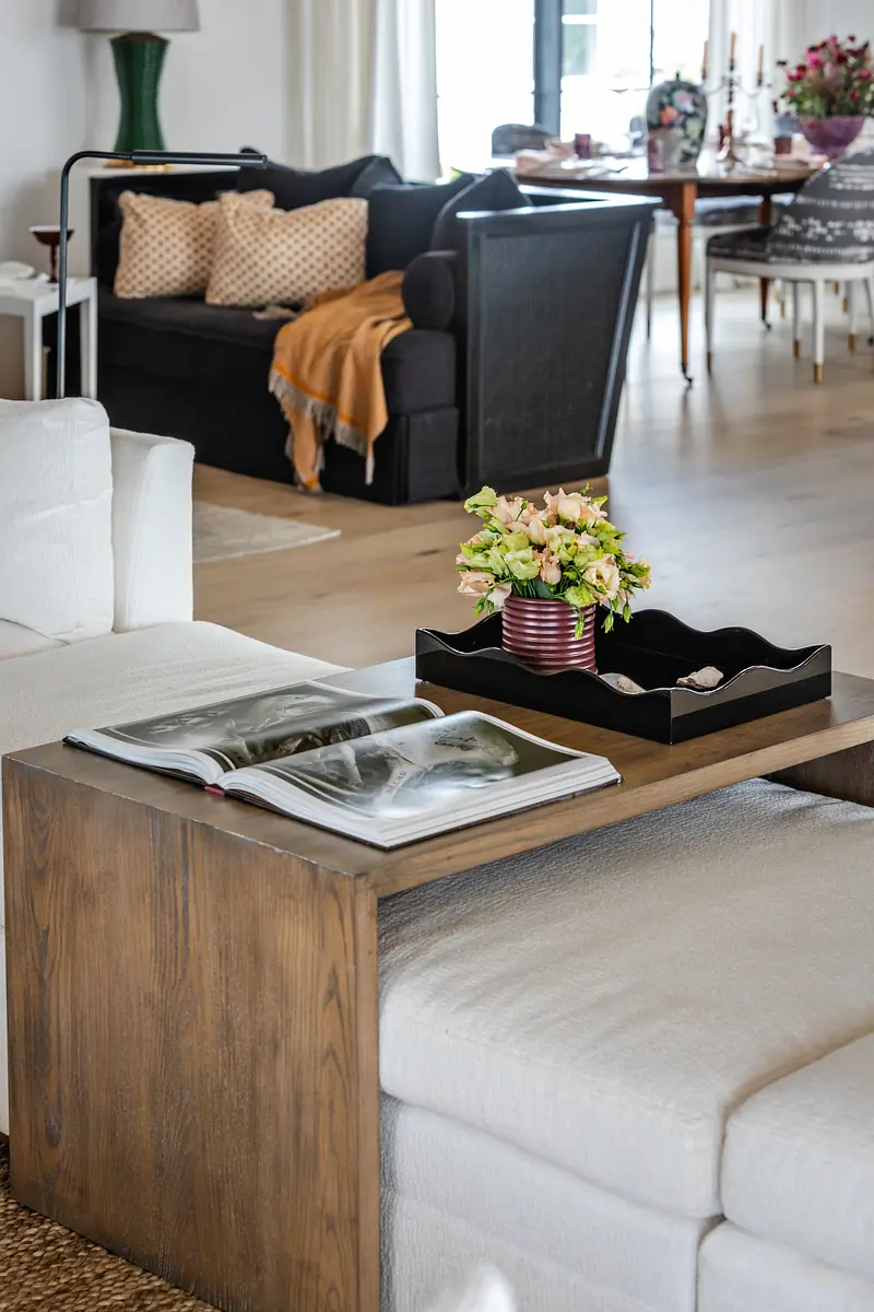 Living room with white sectional, dark accent sofa, wooden side table, flowers, and large windows.