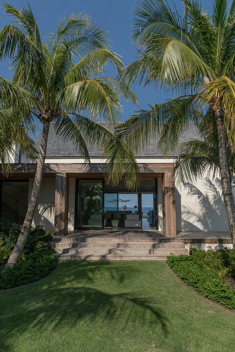 Exterior front view of a modern house with glass doors, wooden framing, and landscaped yard with palm trees.