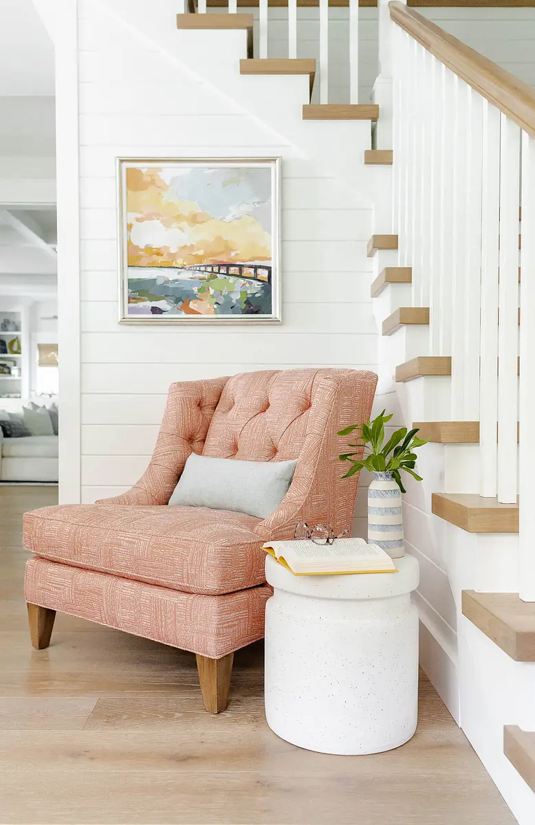 Seating area with coral armchair, white side table, potted plant, artwork, and staircase in the background