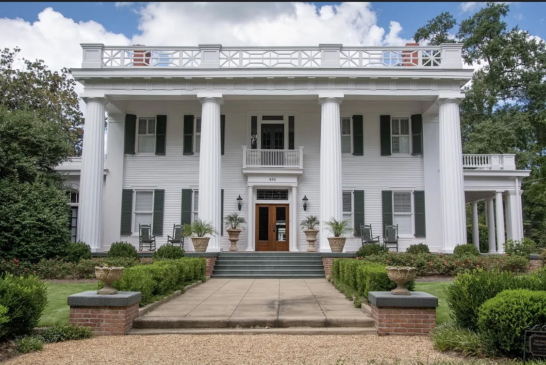 Exterior front view of a two-story house with columns, green shutters, and a large wooden front door.